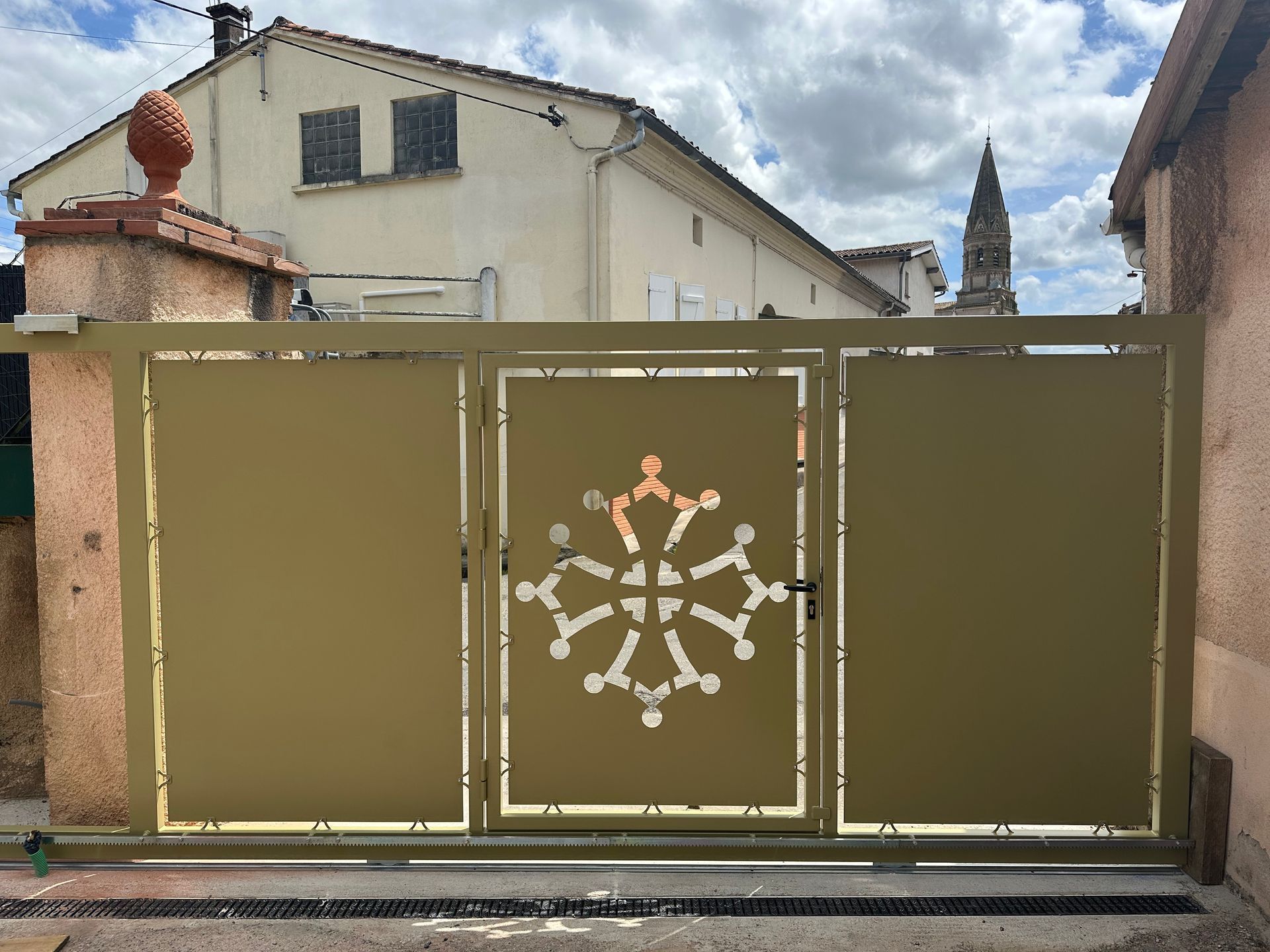 Un portail coulissant en métal beige présente une découpe centrale en forme de croix occitane, installé dans un espace résidentiel extérieur.
