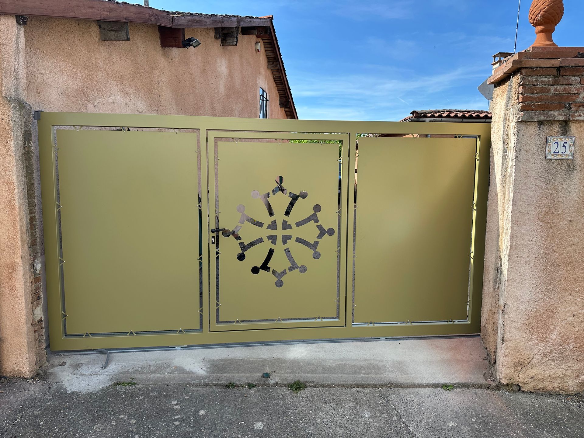 Un portail d'allée en métal doré orné d'une croix occitane découpée en son centre, encadré par deux piliers de pierre.