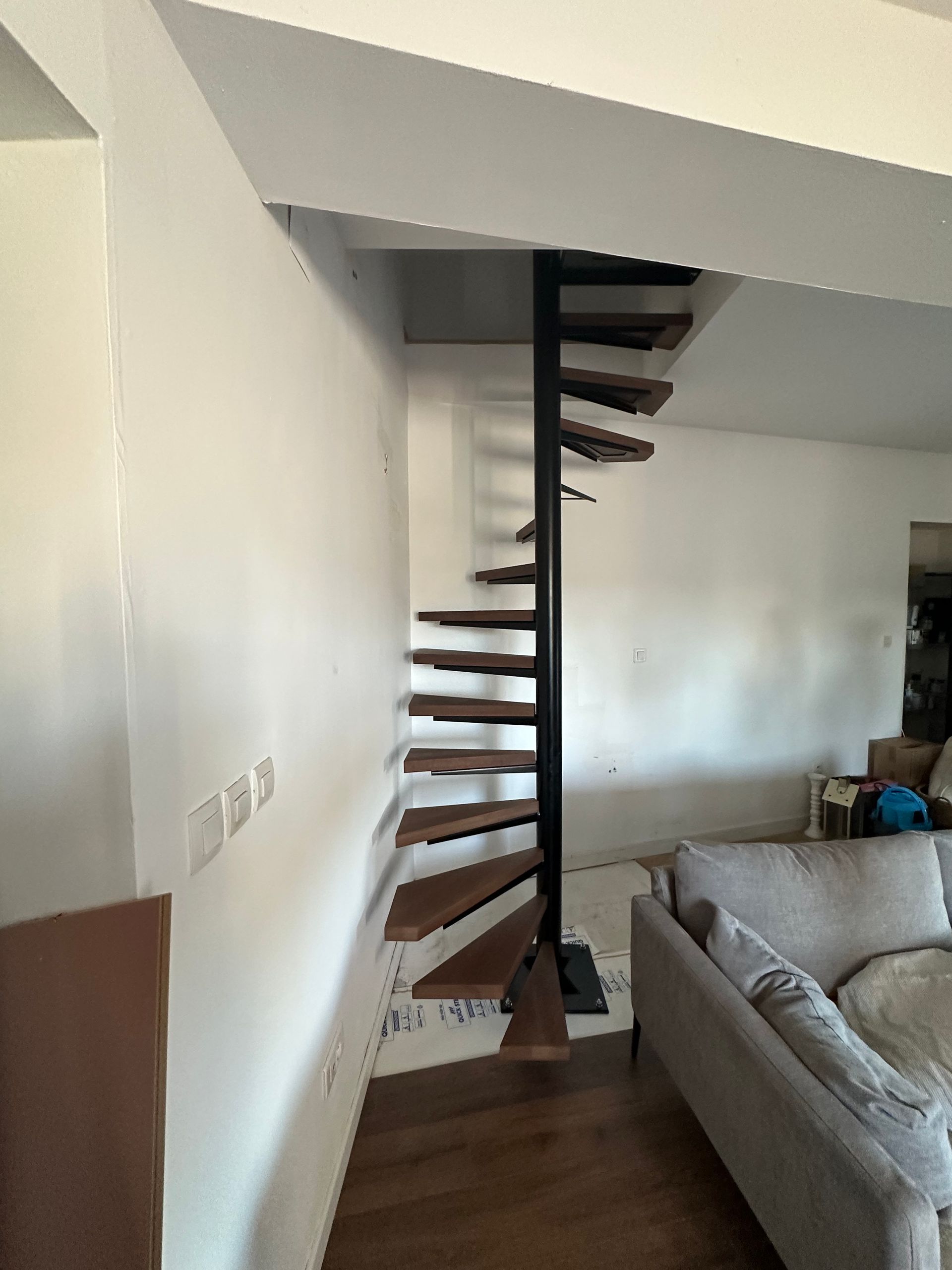 Un escalier en colimaçon avec une colonne centrale en métal noir et des marches en bois, situé dans un salon d'habitation.