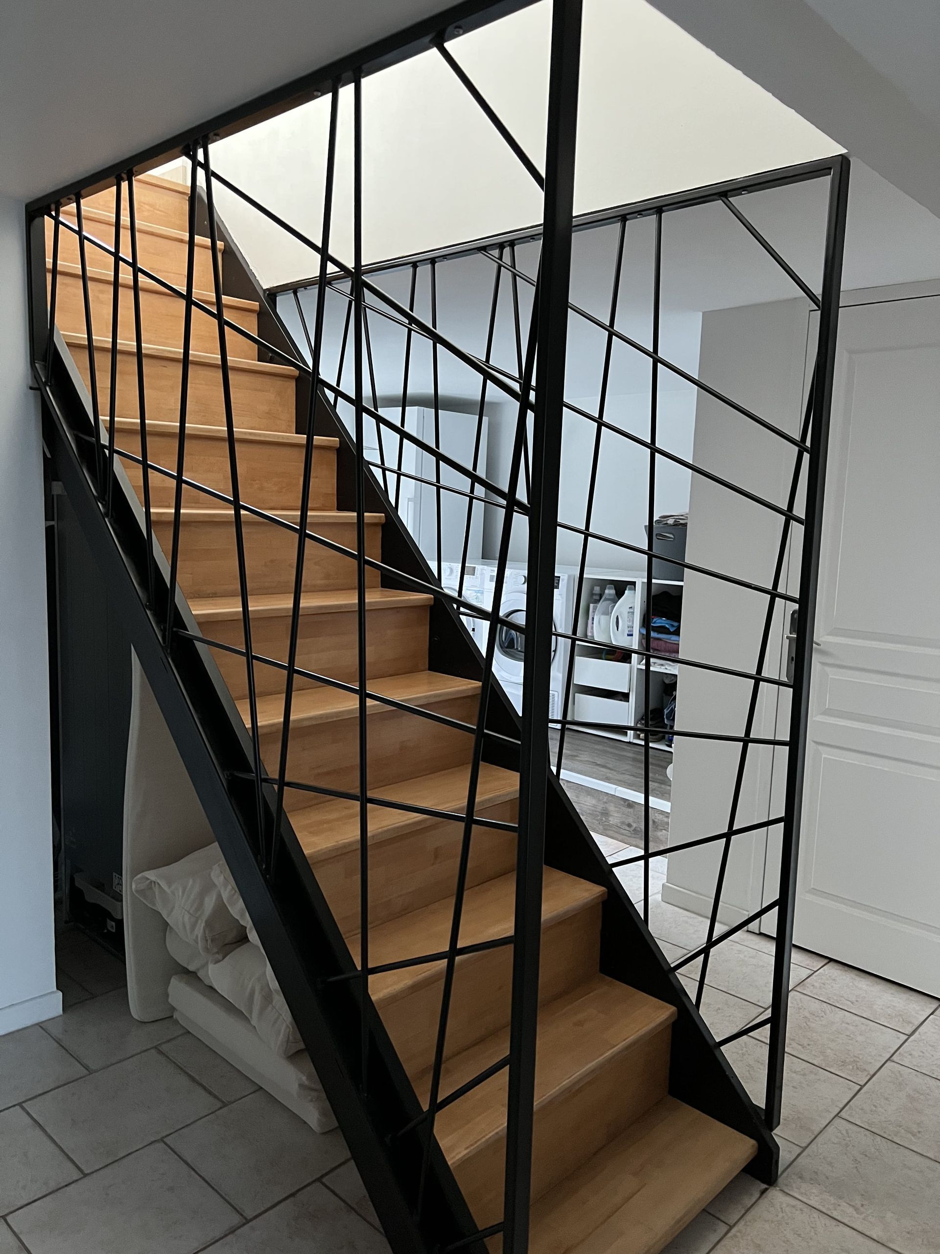 Escalier avec marches en bois clair et structure en métal noir avec une rampe à motif de grille diagonale entrecroisée.