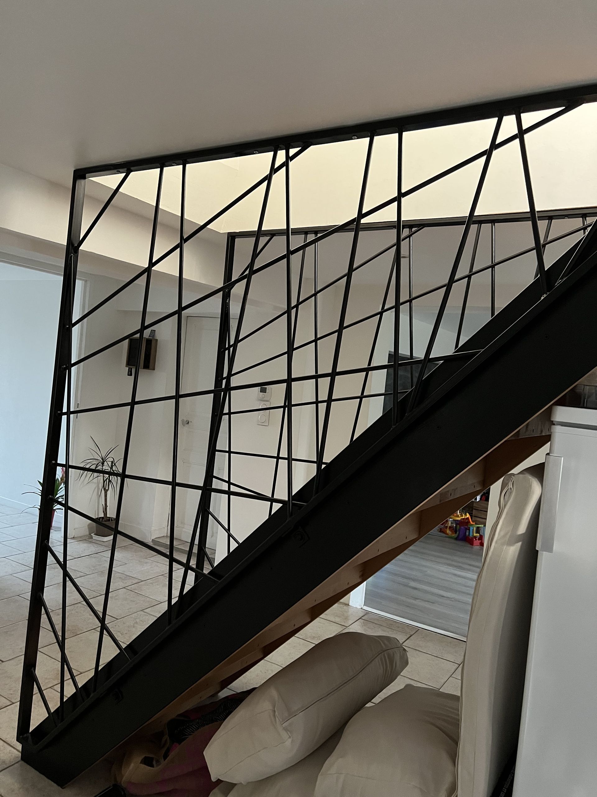 Escalier avec une rampe en métal géométrique noir, partiellement obstrué par des meubles et des coussins dans une pièce.