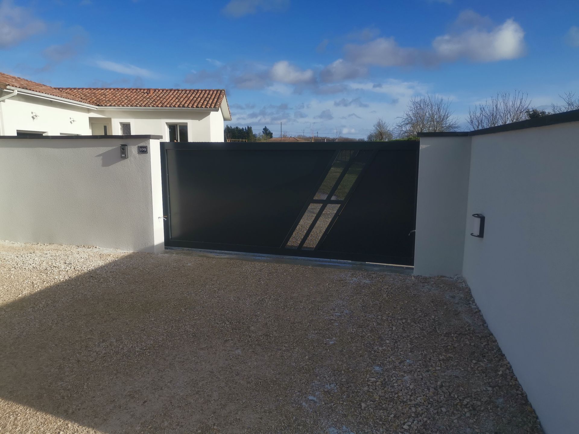 Un portail en métal noir avec deux ouvertures diagonales pour les fenêtres, encadré par des murs en stuc blanc et une allée de gravier.