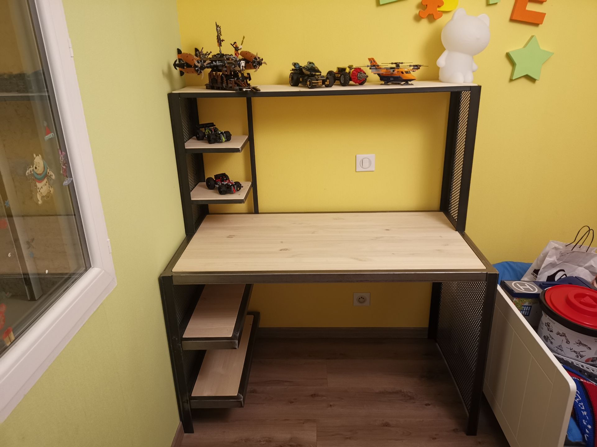 Un bureau aux surfaces en bois clair, à la structure en métal foncé et aux étagères latérales ouvertes, placé contre un mur jaune dans une pièce.