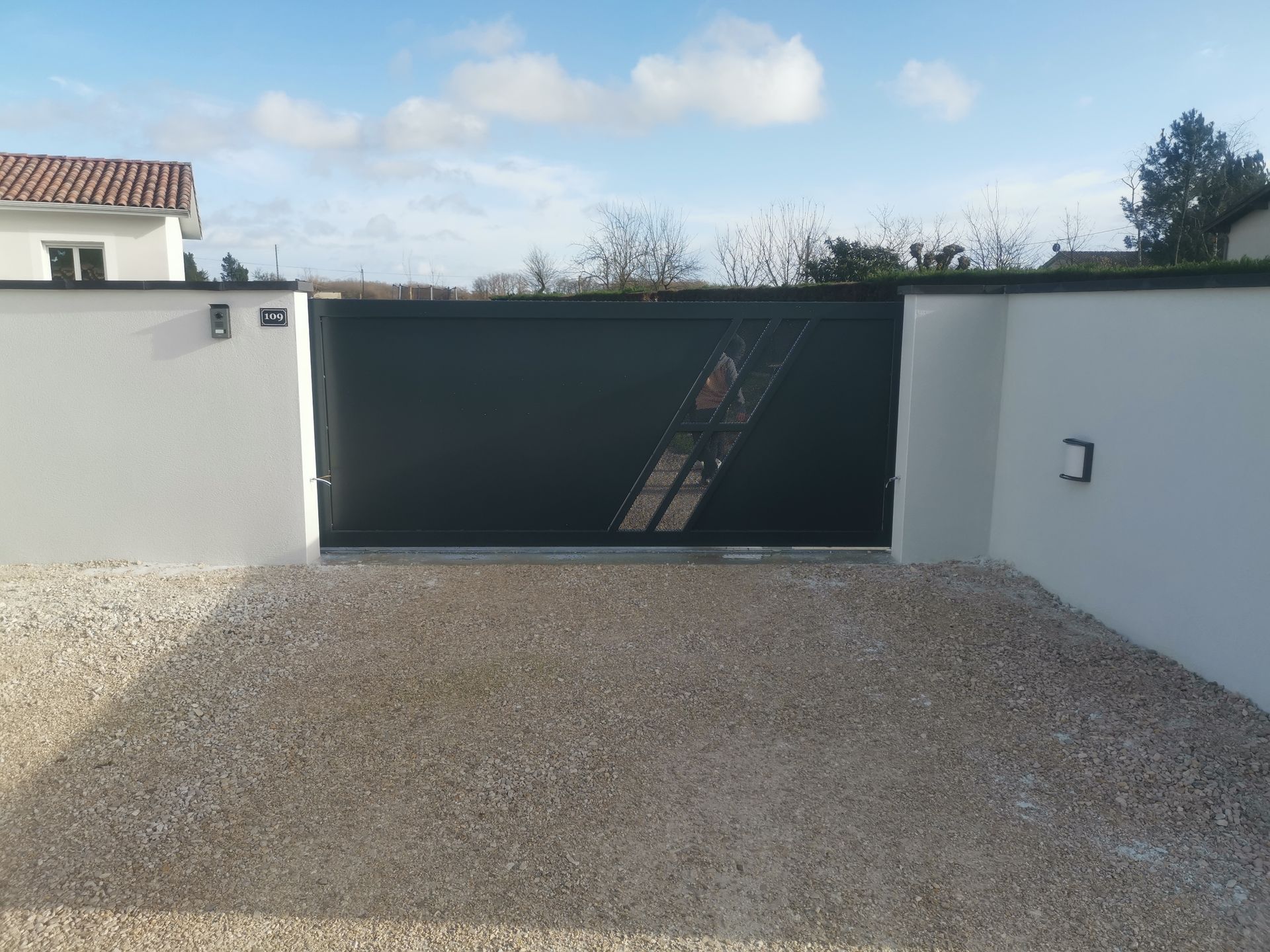 Un portail d'allée moderne, gris foncé, orné d'une découpe décorative en diagonale, situé entre des murs blancs sur un terrain gravillonné.