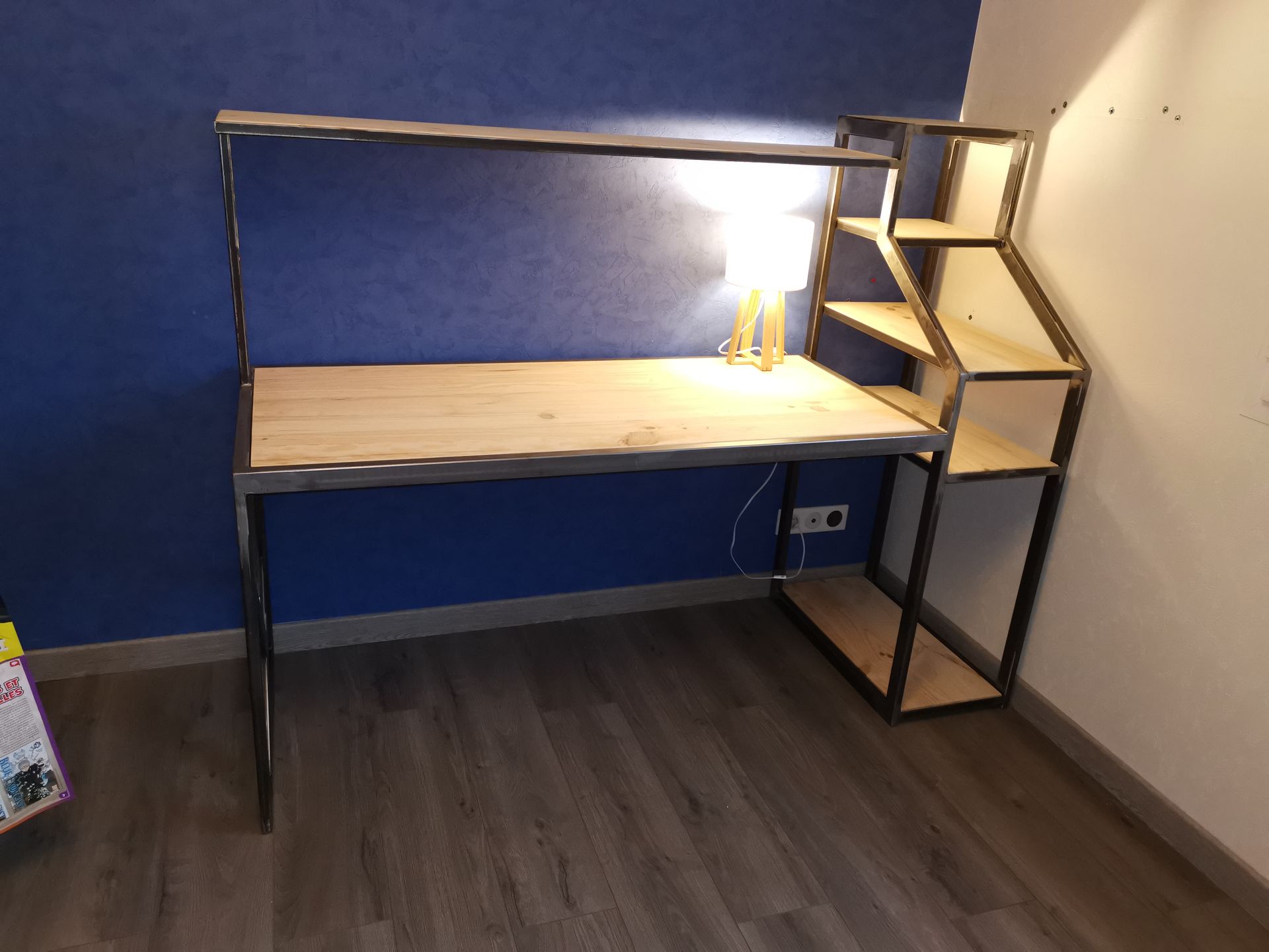 Un bureau en bois avec une structure en métal noir et des étagères intégrées, adossé à un mur bleu surmonté d'une petite lampe.