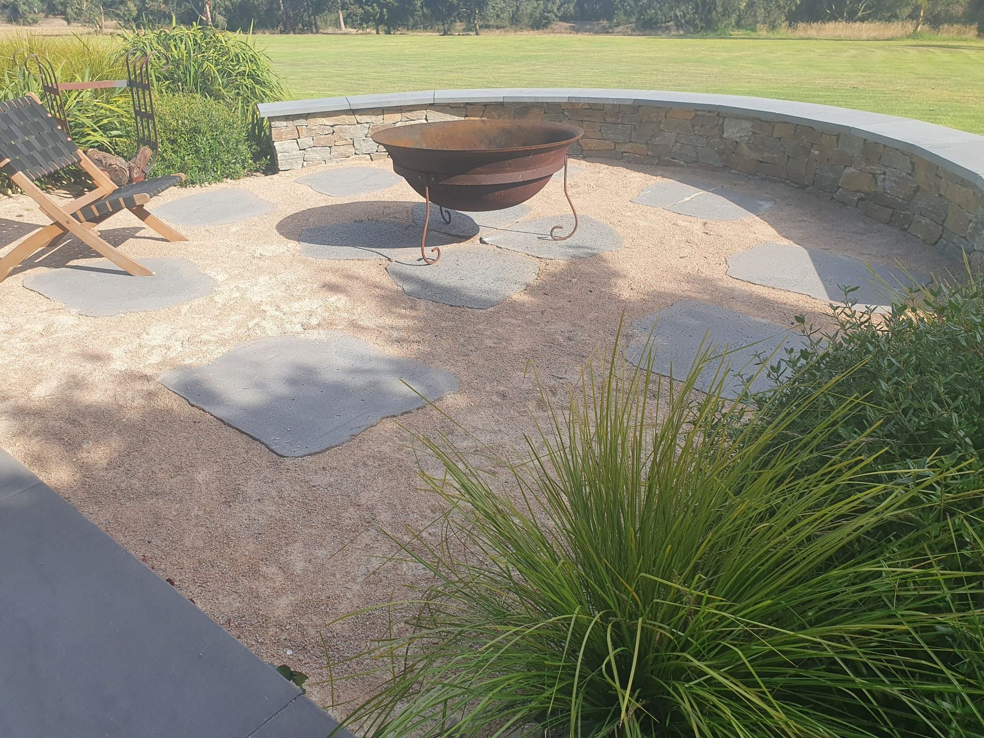 Outdoor patio with stone fire pit, chairs, and gravel, surrounded by curved stone wall and lawn.