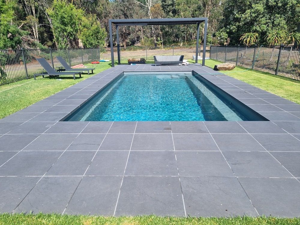 Rectangular pool with dark gray tile surround and patio furniture in a grassy yard, trees in the background.