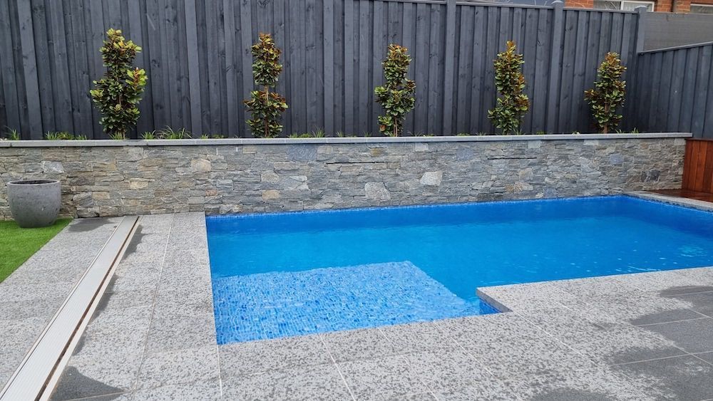 Pool with blue water and tiled steps, surrounded by stone and gray patio. Trees in background.
