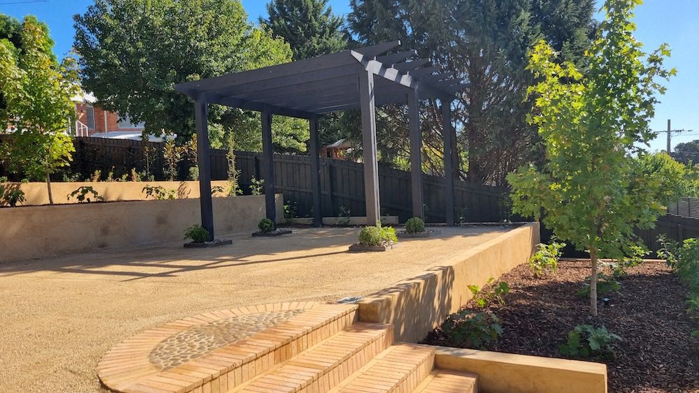 Pergola with gravel path and brick steps in a landscaped garden with trees and retaining walls.