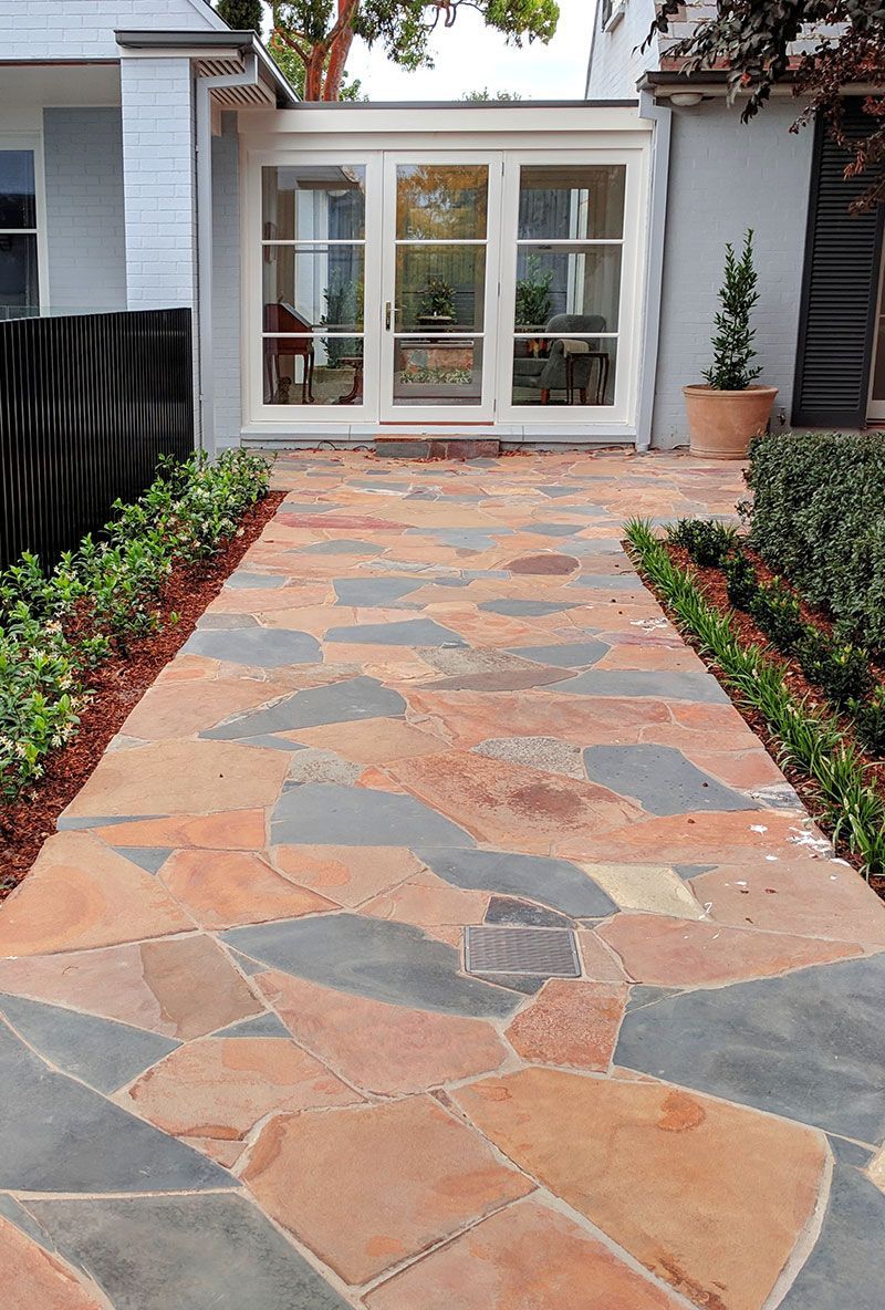 Stone path leading to a white-framed glass enclosure; bordered by green hedges and flowerbeds.