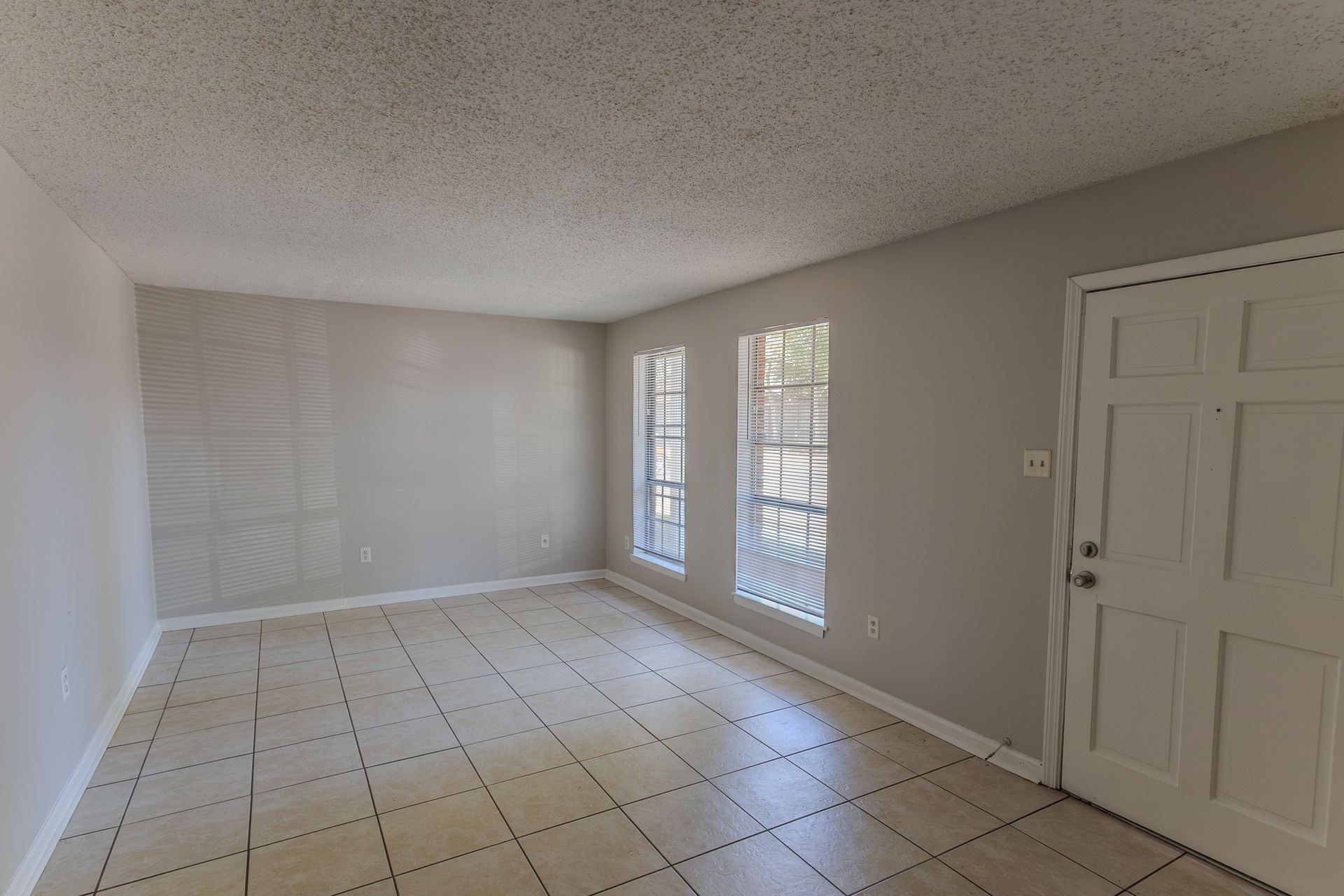 Empty, gray-painted room with tile flooring, two windows with bars, and a white door.