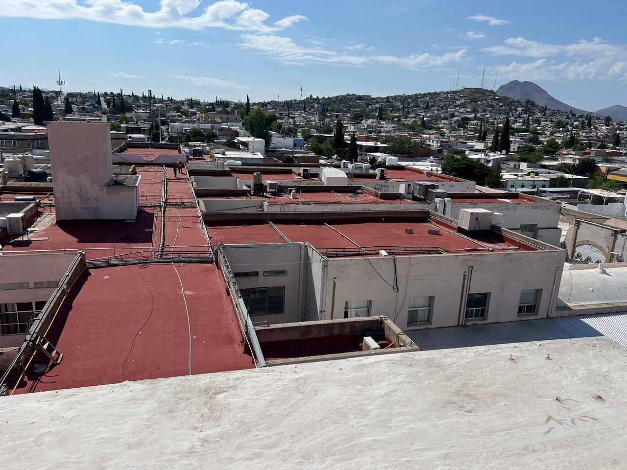 Vista aérea de un complejo de edificios con techo rojo en un área urbana con un fondo montañoso y un cielo azul.