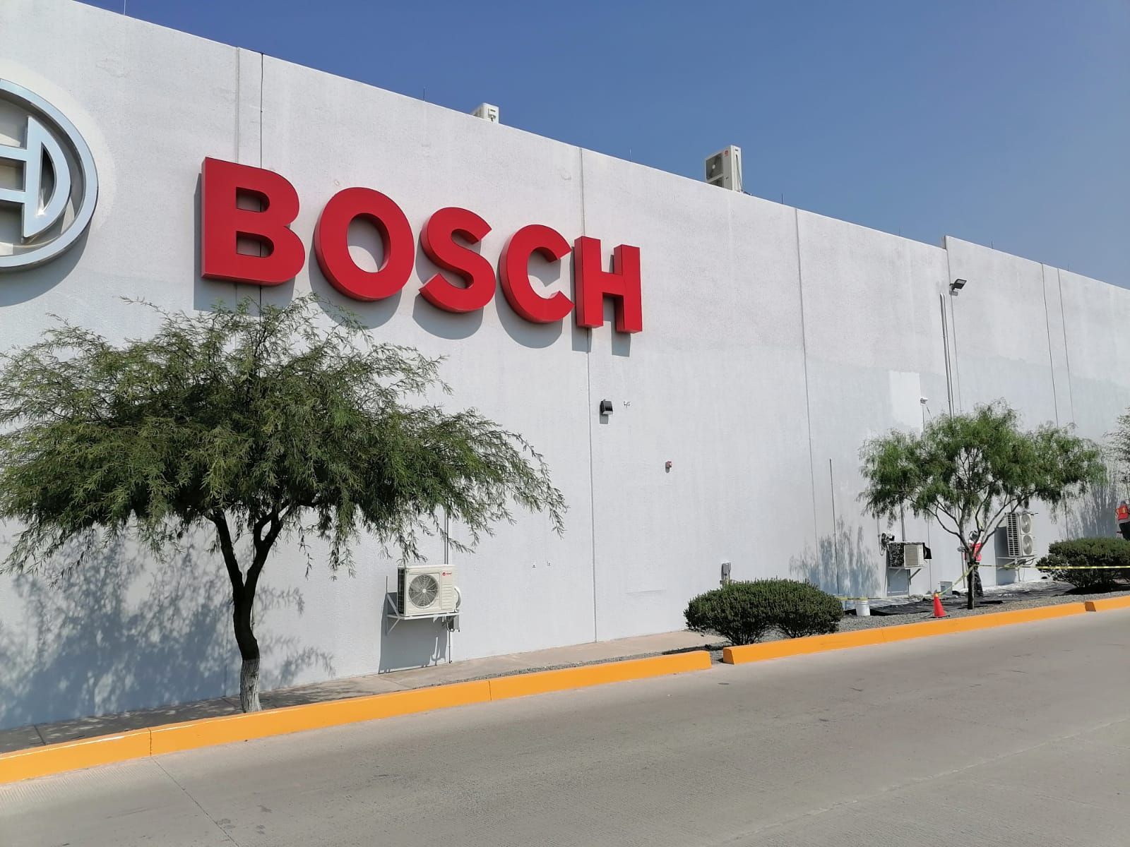 Exterior de la fábrica de Bosch con logotipo rojo sobre un edificio blanco, árboles y calle al frente.
