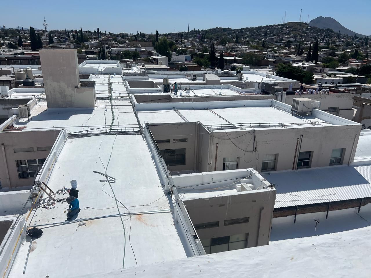 Vista aérea de un tejado blanco y plano con una persona trabajando. Al fondo, edificios y un paisaje montañoso.