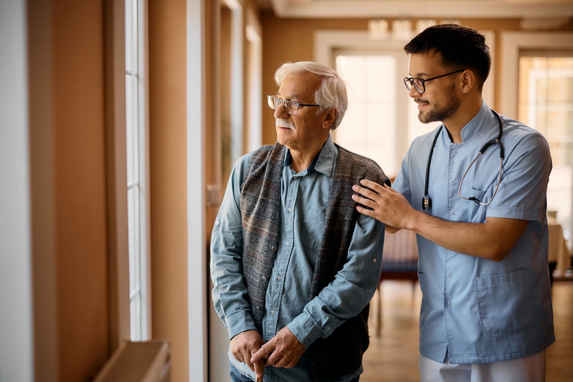 caretaker helping elderly man walk, as pair looks out window