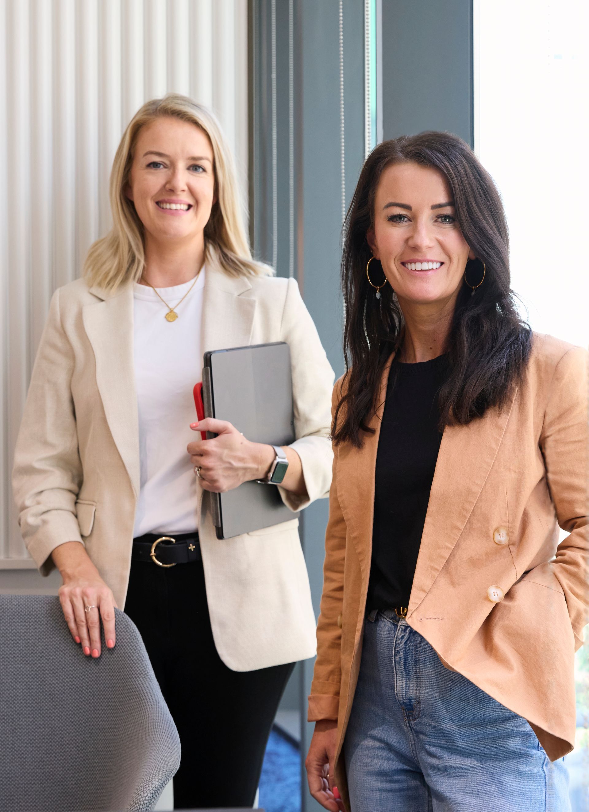 Two women are standing next to each other in a room and smiling . one of the women is holding a laptop.