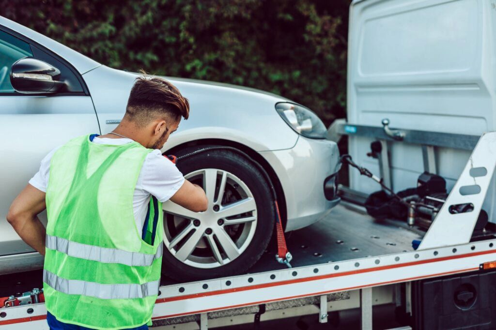 Man in Safety Vest Securing Silver Car Onto a Tow Truck on a Road — Aura Towing Service in Landsborough, QLD 