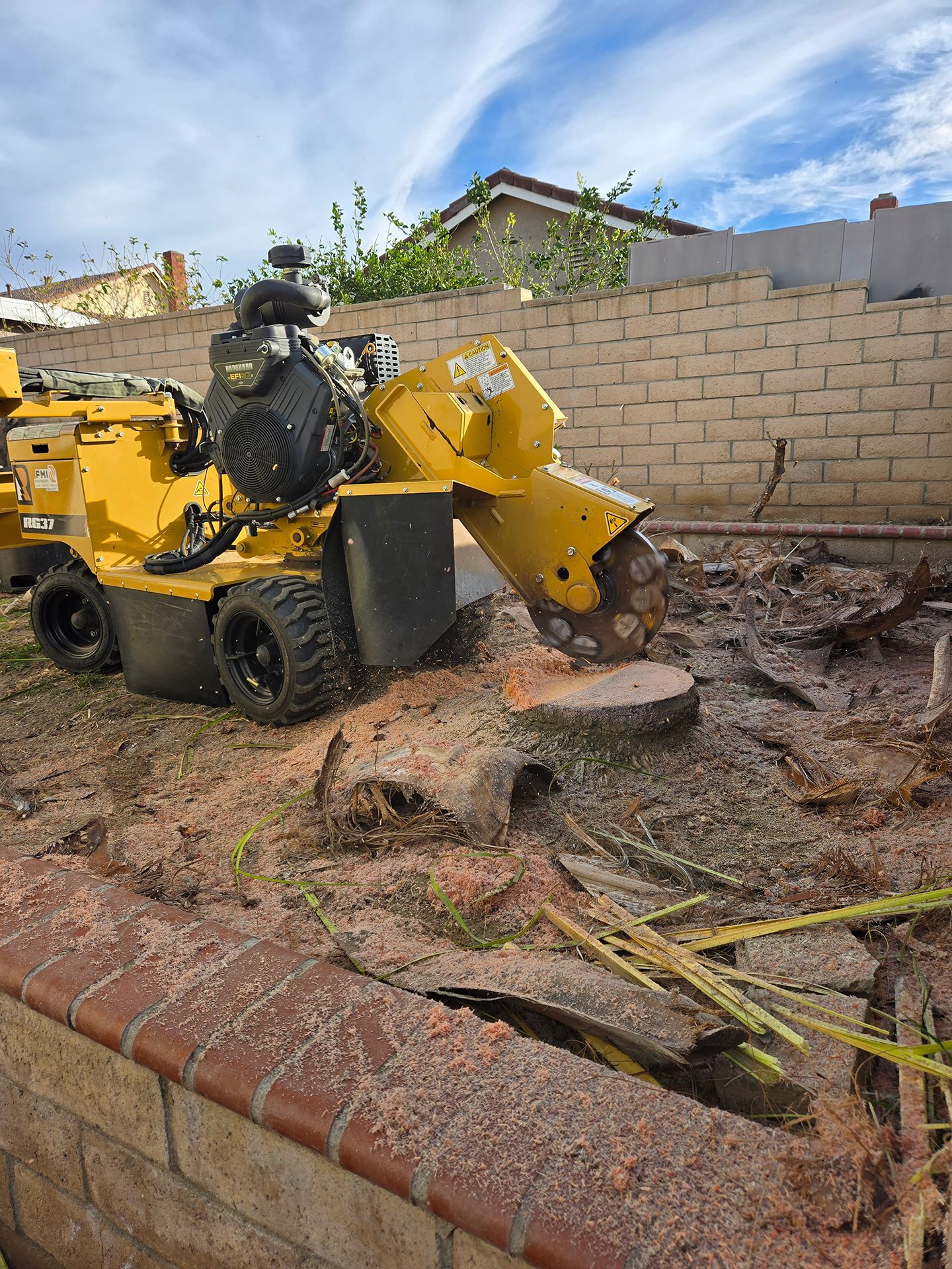 A yellow stump grinder is cutting a tree stump in front of a brick wall