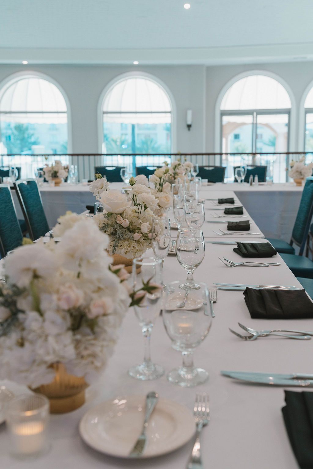 Elegant banquet hall with long white-tablecloth tables, floral centerpieces, and arched windows overlooking water