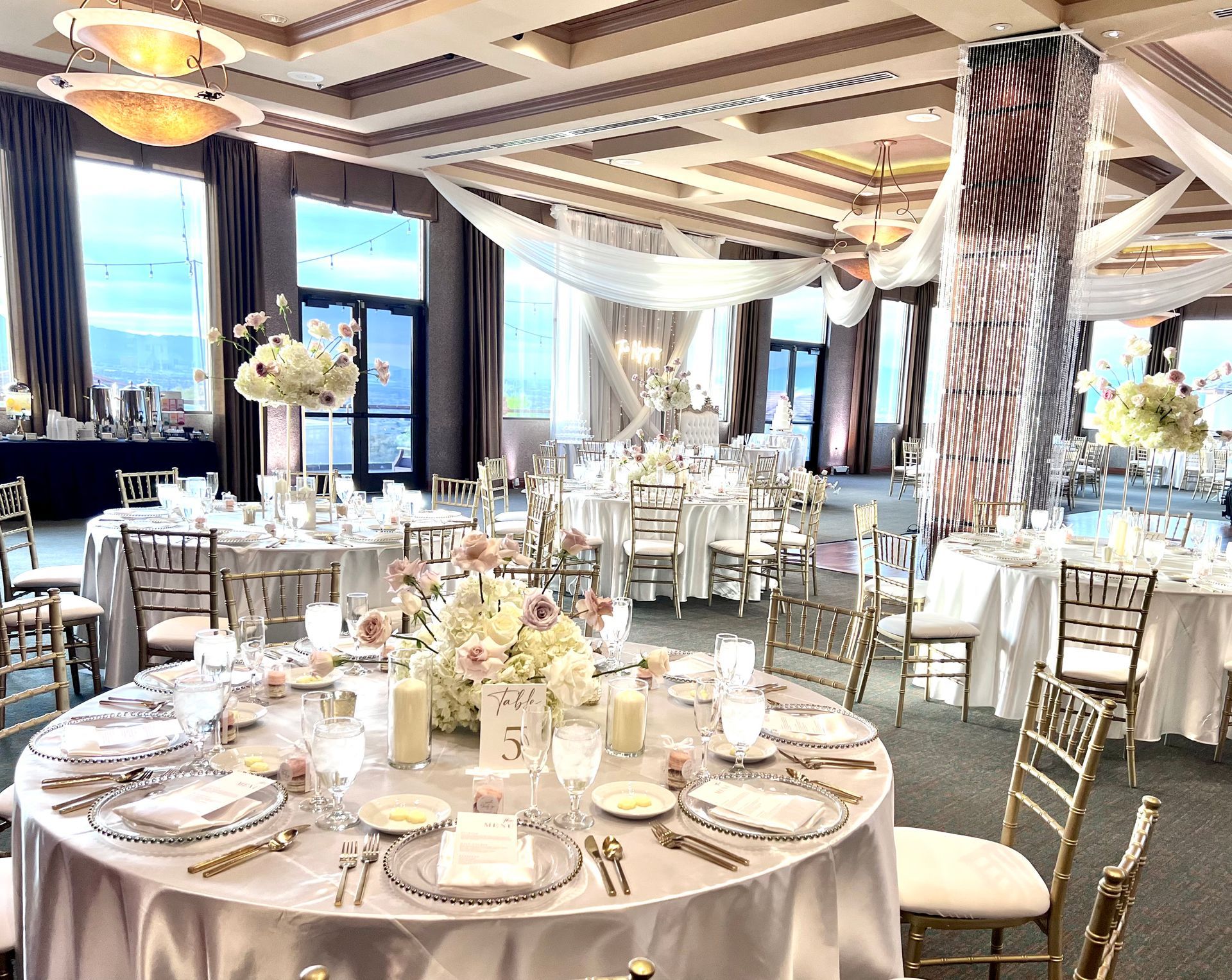 Elegant banquet hall with round, flower-decorated tables, white chairs, and a city view through large windows