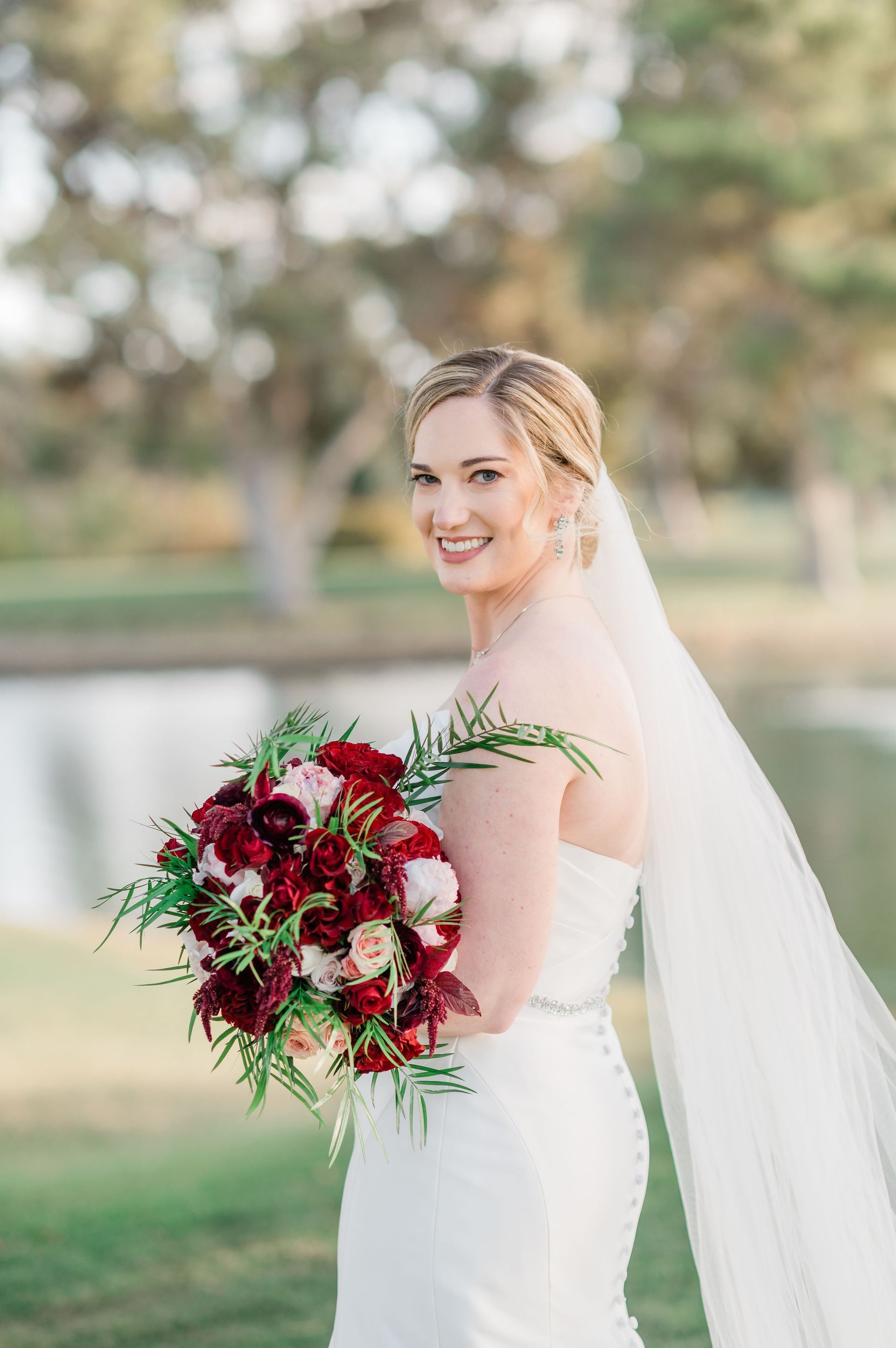 Bride in a white dress holding a red bouquet outdoors, smiling with a long veil.