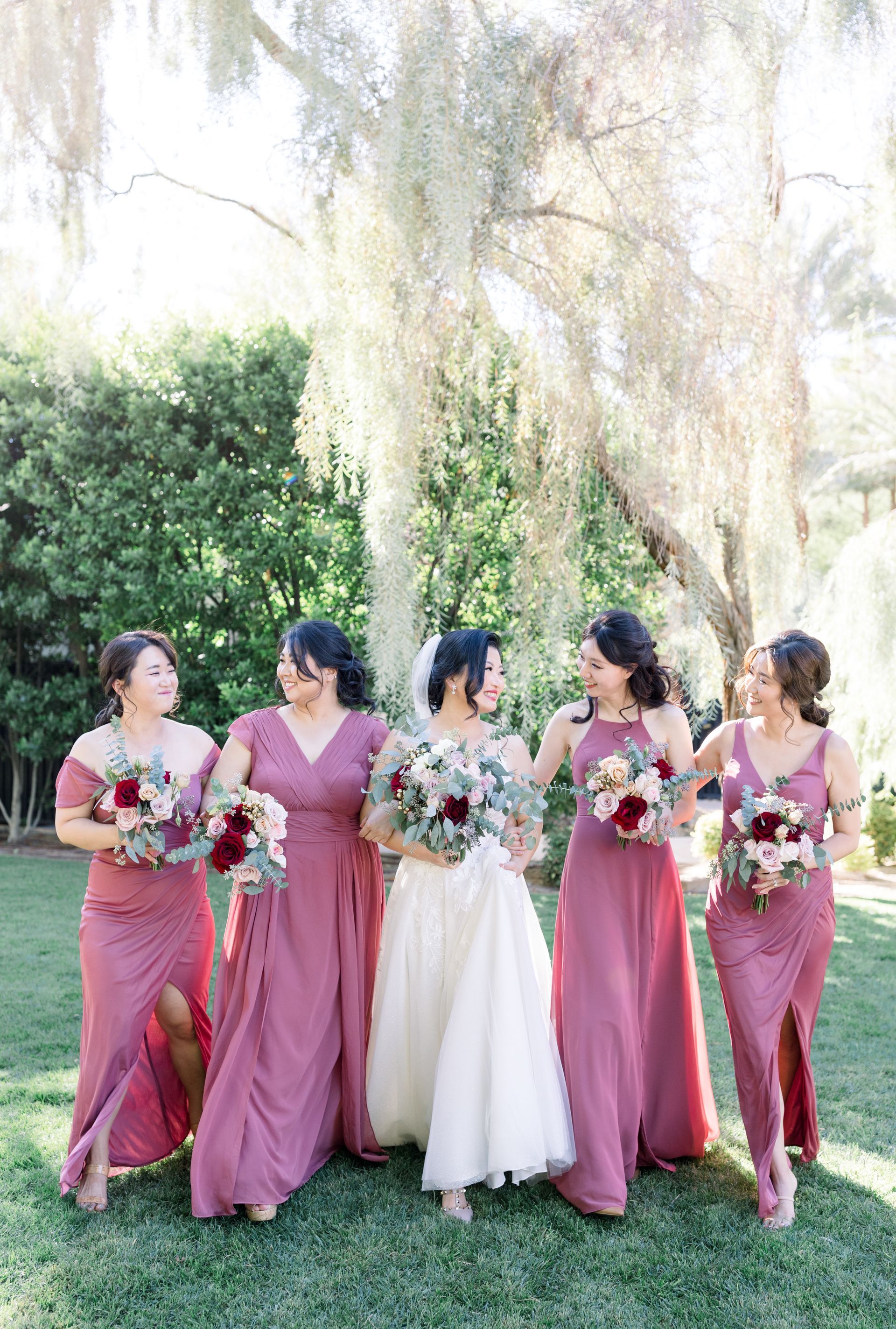 Five women in pink and white dresses holding bouquets outdoors under hanging trees.
