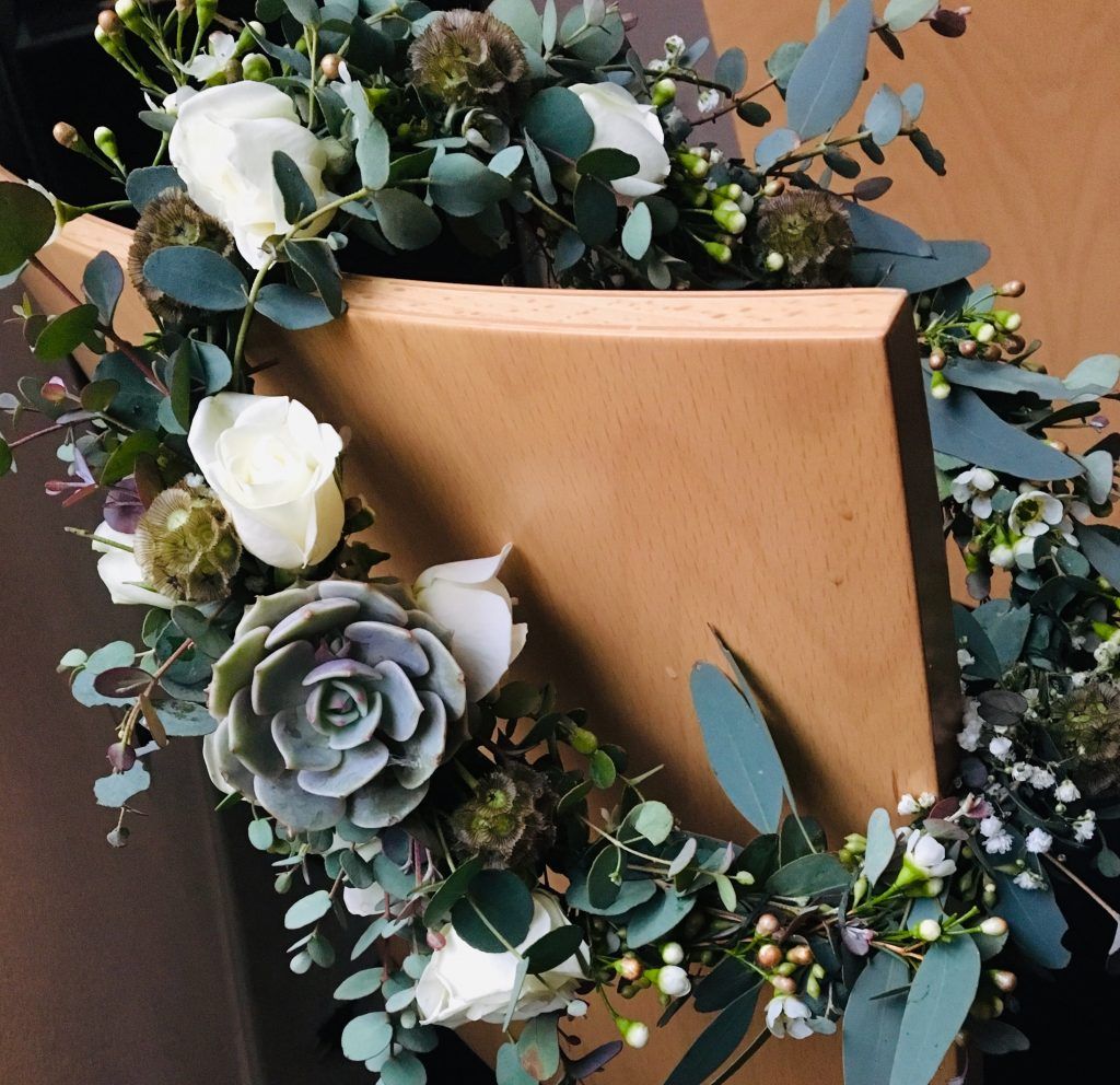 Wooden plaque framed by a lush wreath of white flowers, eucalyptus, and a gray succulent.