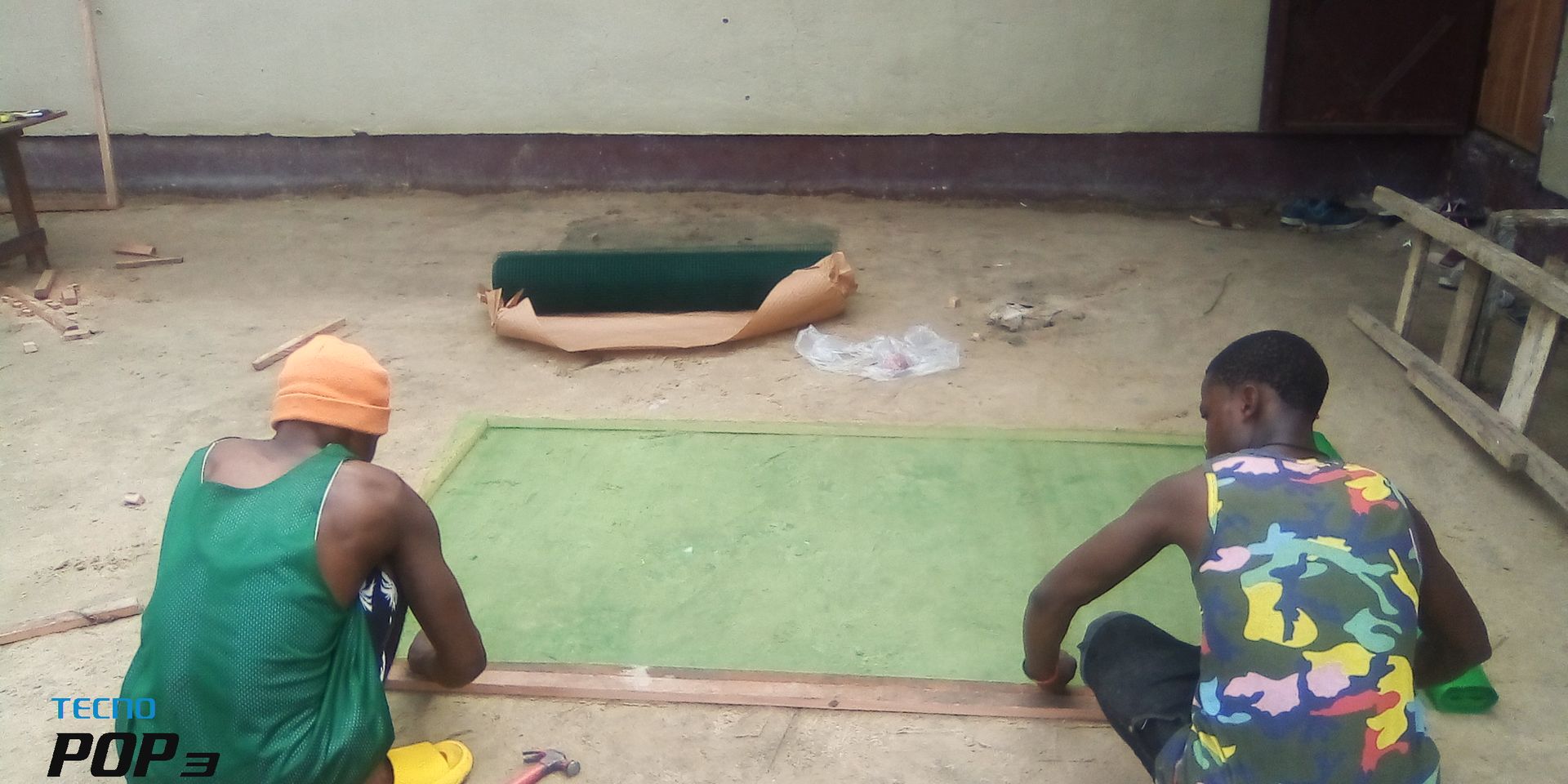 Two people crouch on a floor beside a green mat, with another person lying face down near the wall.
