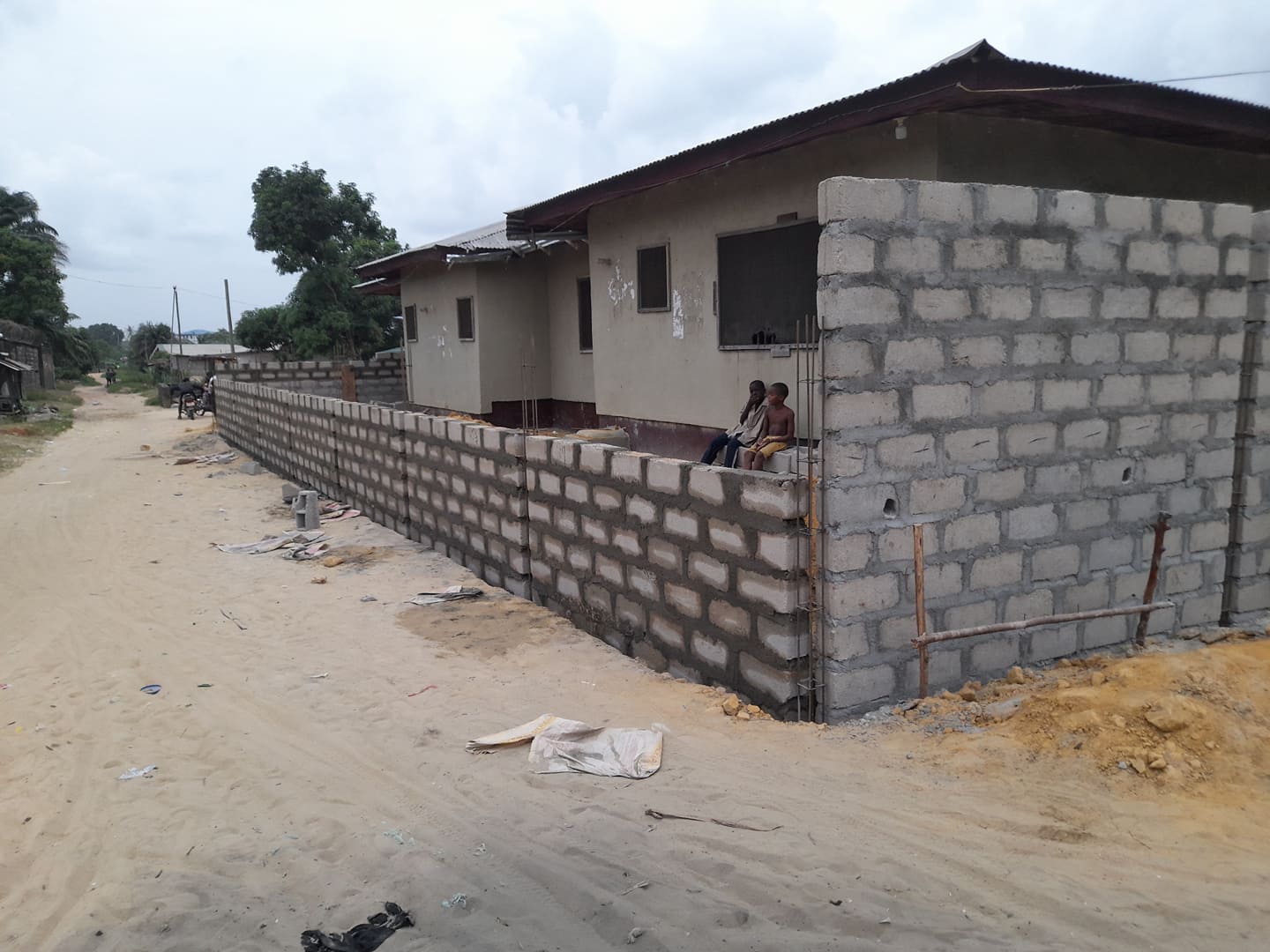 Partially built cinderblock house along a sandy road, with two people seated by the wall.