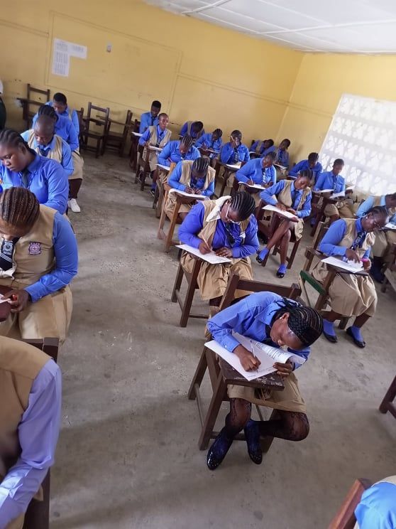 Students in blue uniforms taking a written exam in a classroom, seated at wooden desks.