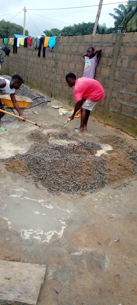 People spreading wet gravel on a street beside a brick wall, with laundry hanging nearby