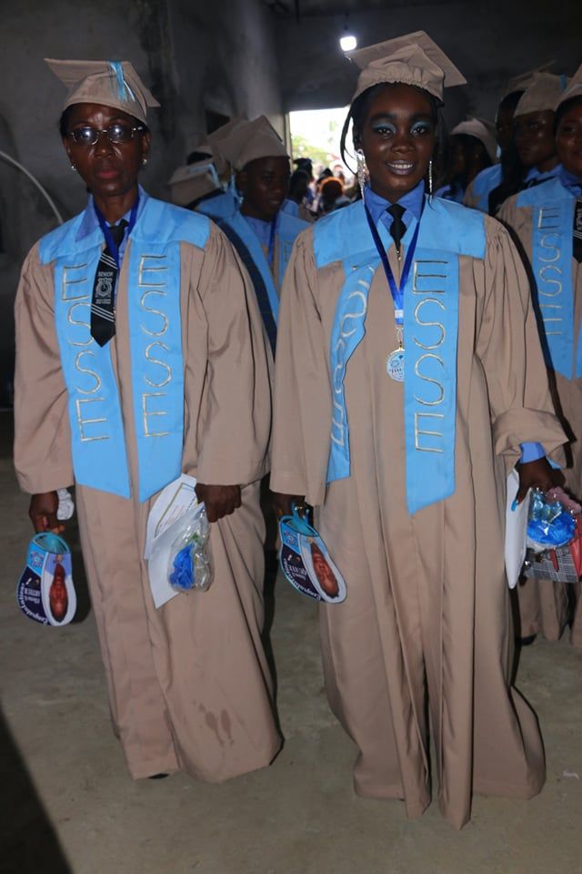 Two people in tan traditional outfits and blue sashes standing outdoors at night during a ceremony