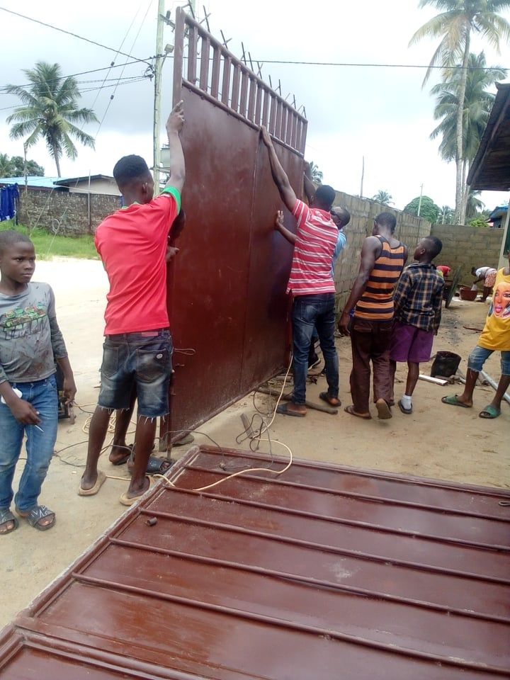 Several people lift a brown metal gate upright near a road, with a child watching nearby.
