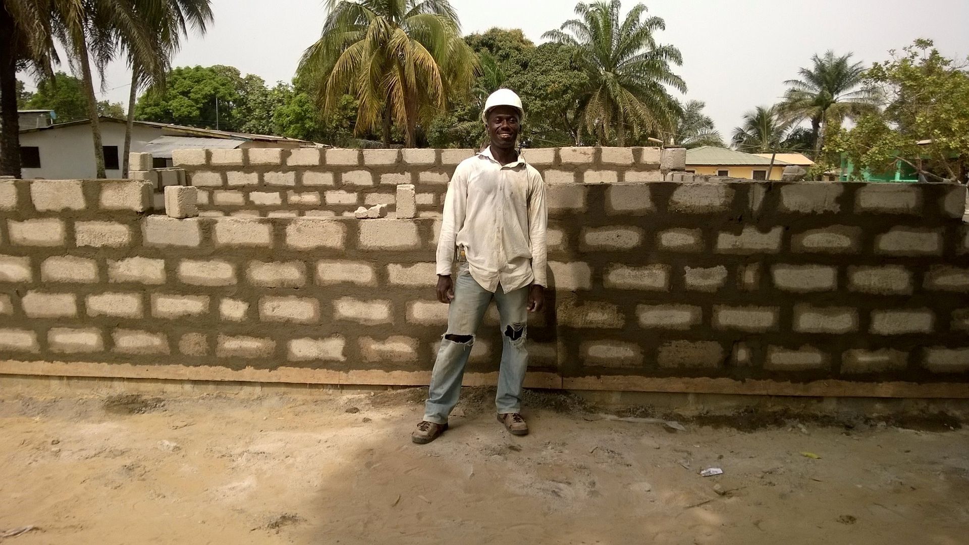 Person standing in front of a low brick wall in a sandy yard with palm trees behind.