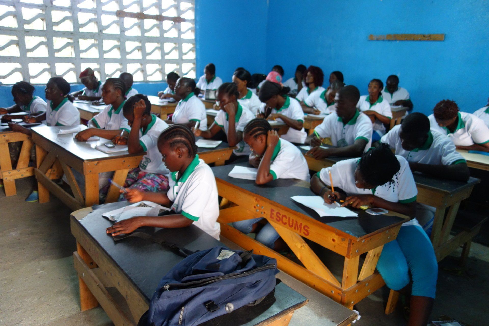 Students in a classroom writing at desks beside bright blue walls and a patterned window