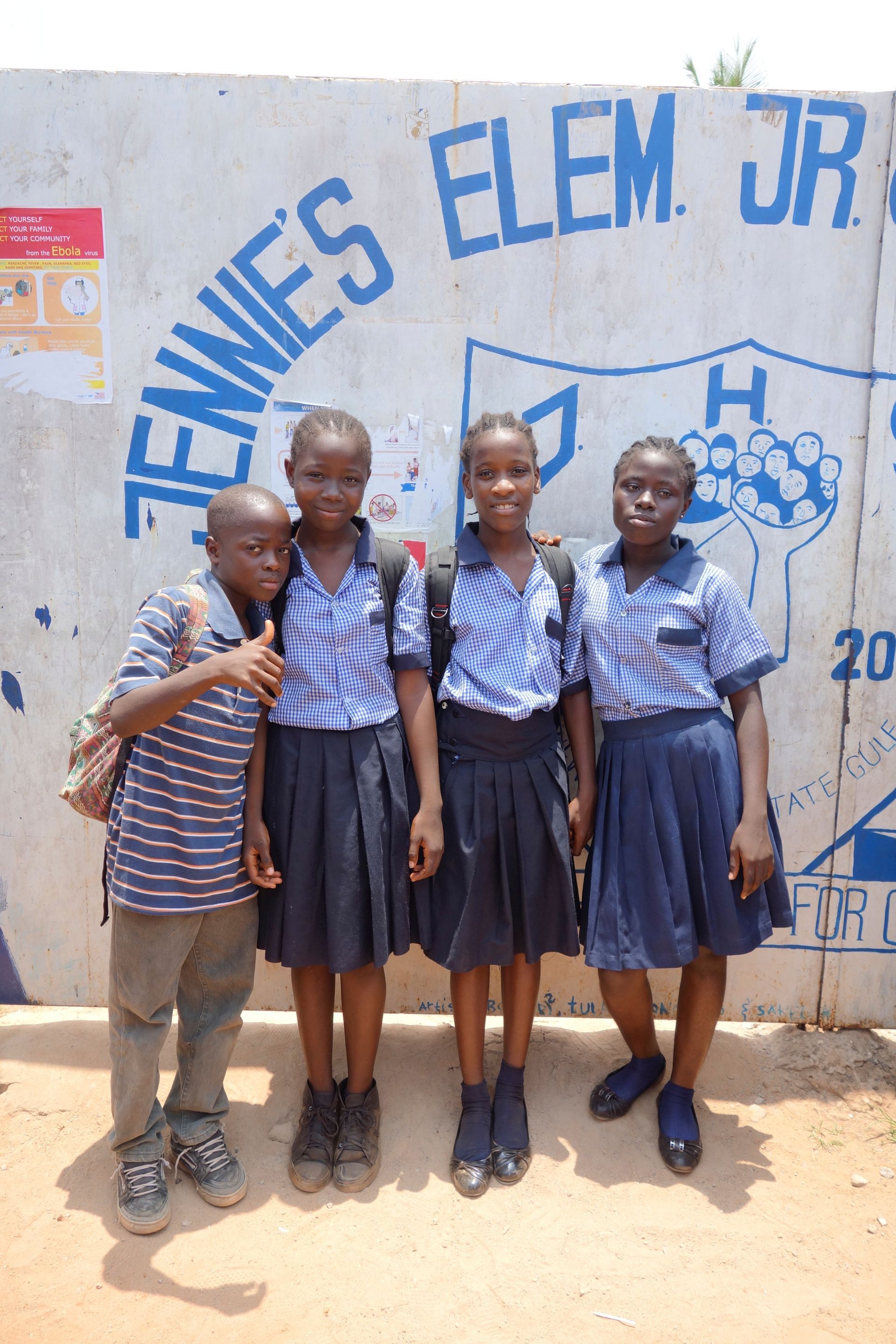 Four schoolchildren in blue uniforms pose outside a wall painted “ENNIES ELEM. JR.”