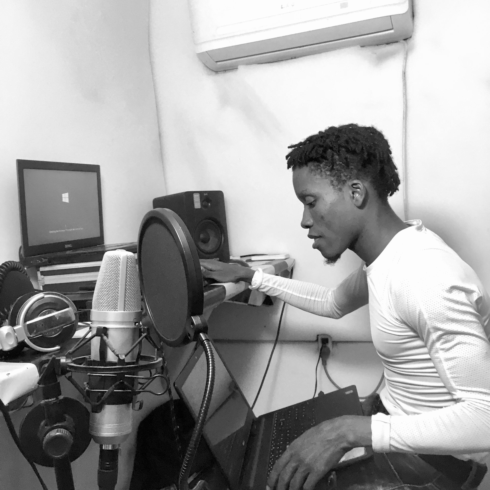 Person recording audio at a desk with microphone, pop filter, laptop, and speakers in a small studio.