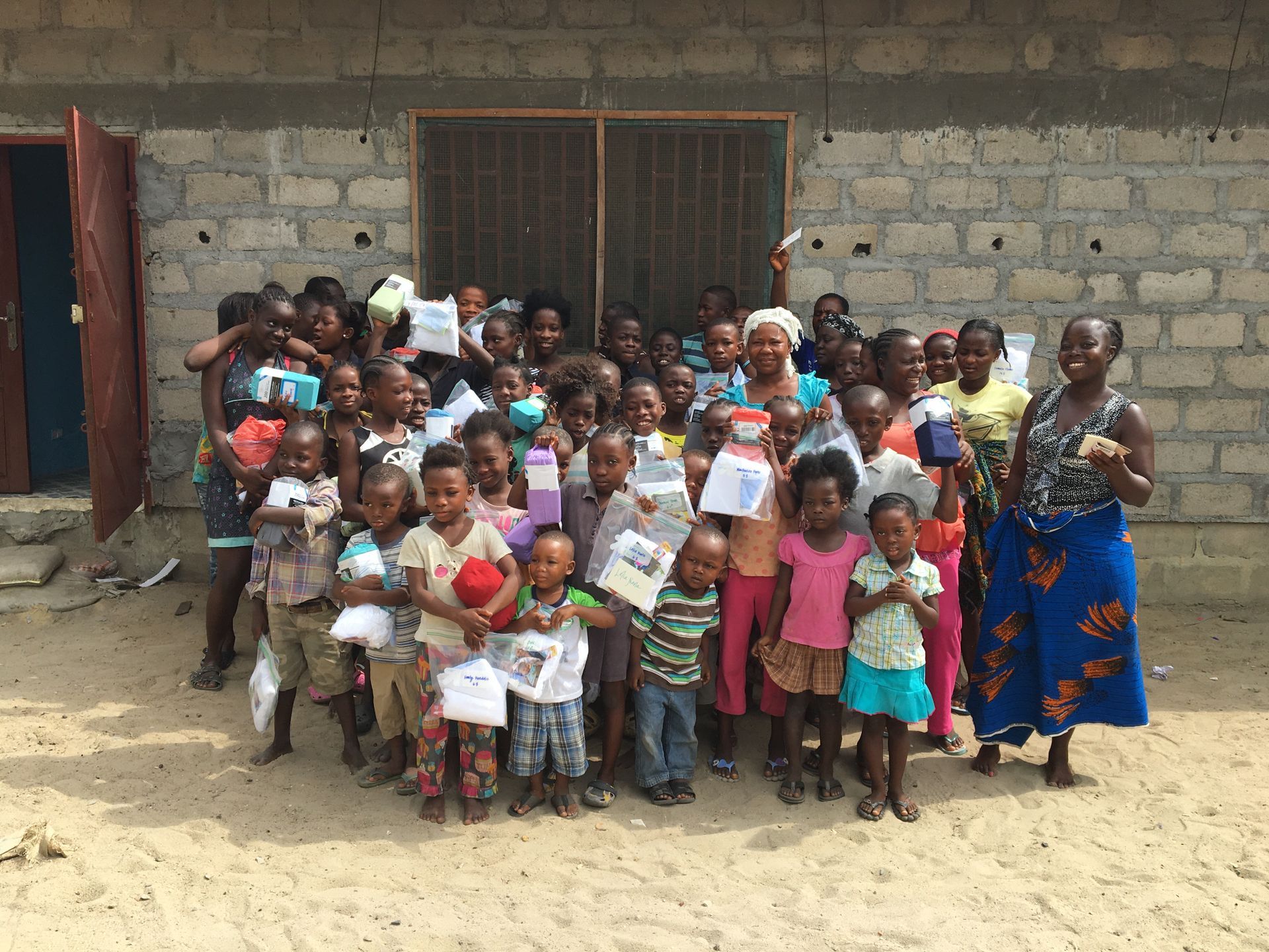 Group of children and a woman standing outside a brick building, many holding papers.