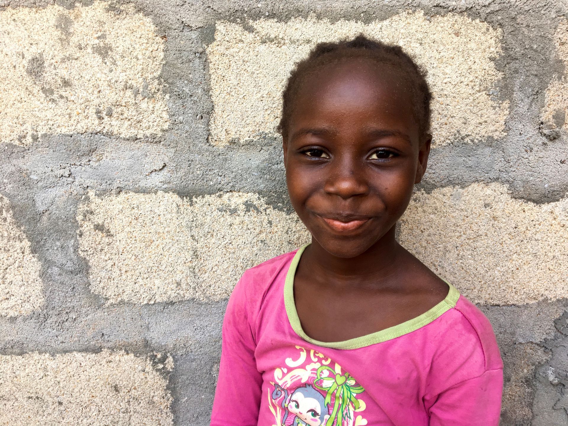 Smiling child in a pink shirt standing against a stone wall