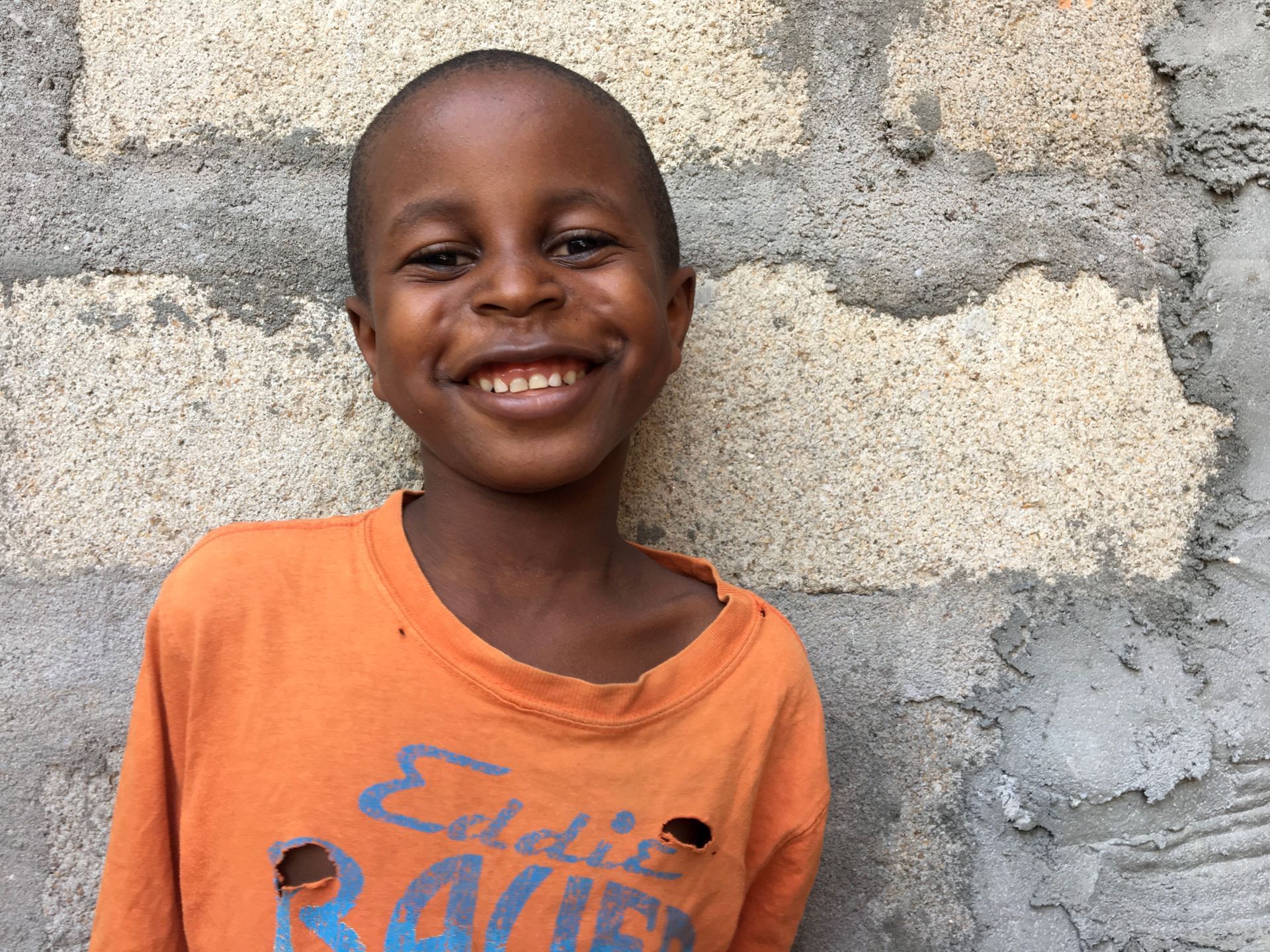 Smiling child in an orange shirt standing against a gray concrete wall.