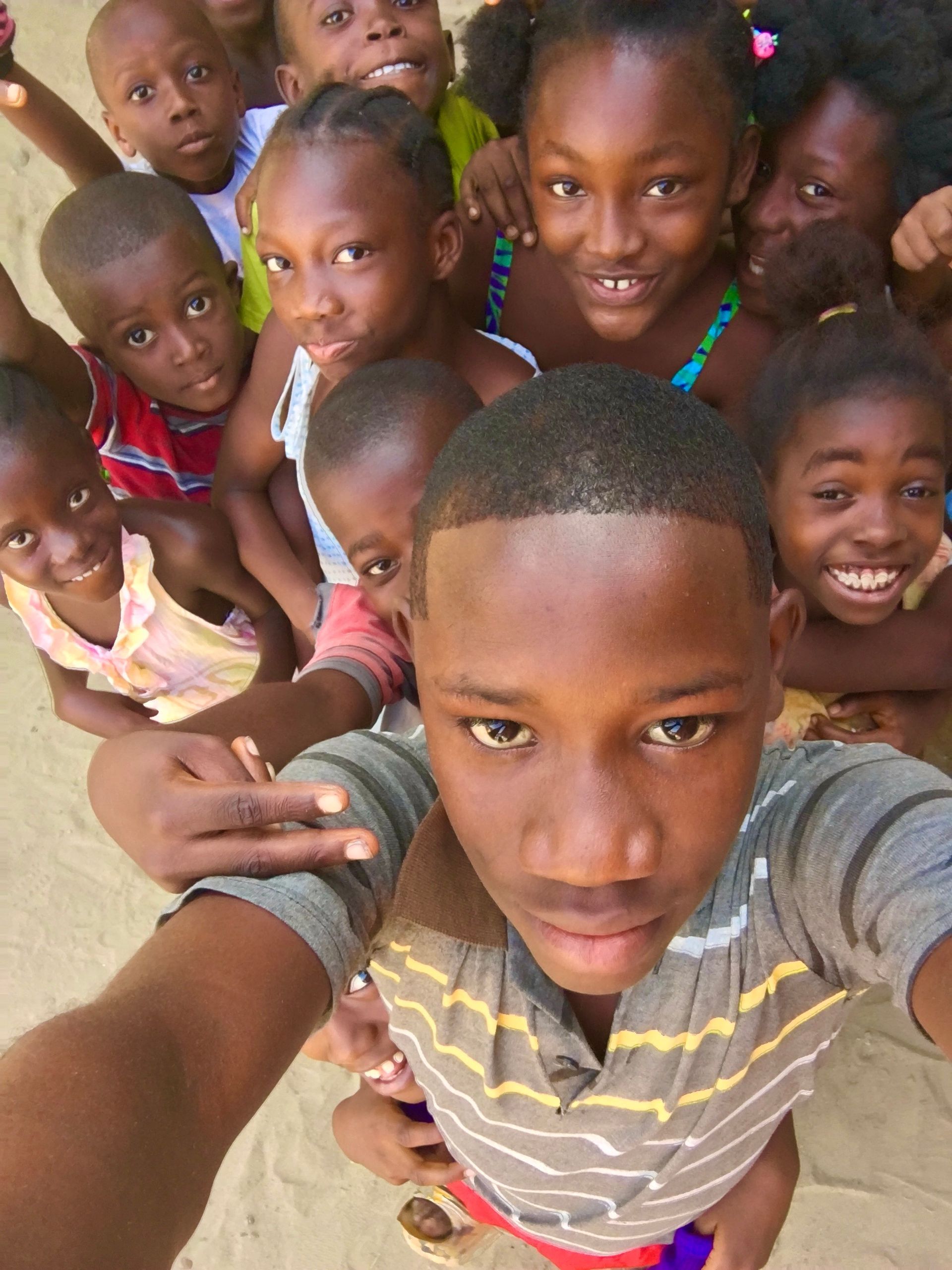 Group of smiling children crowd together for a selfie outdoors on a sandy ground.