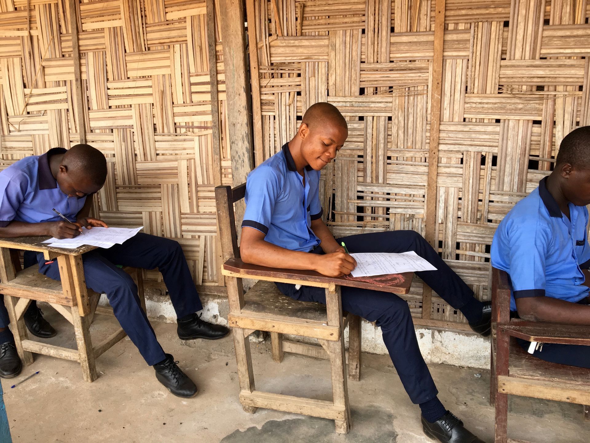 Students in blue uniforms writing at desks against a woven wall background