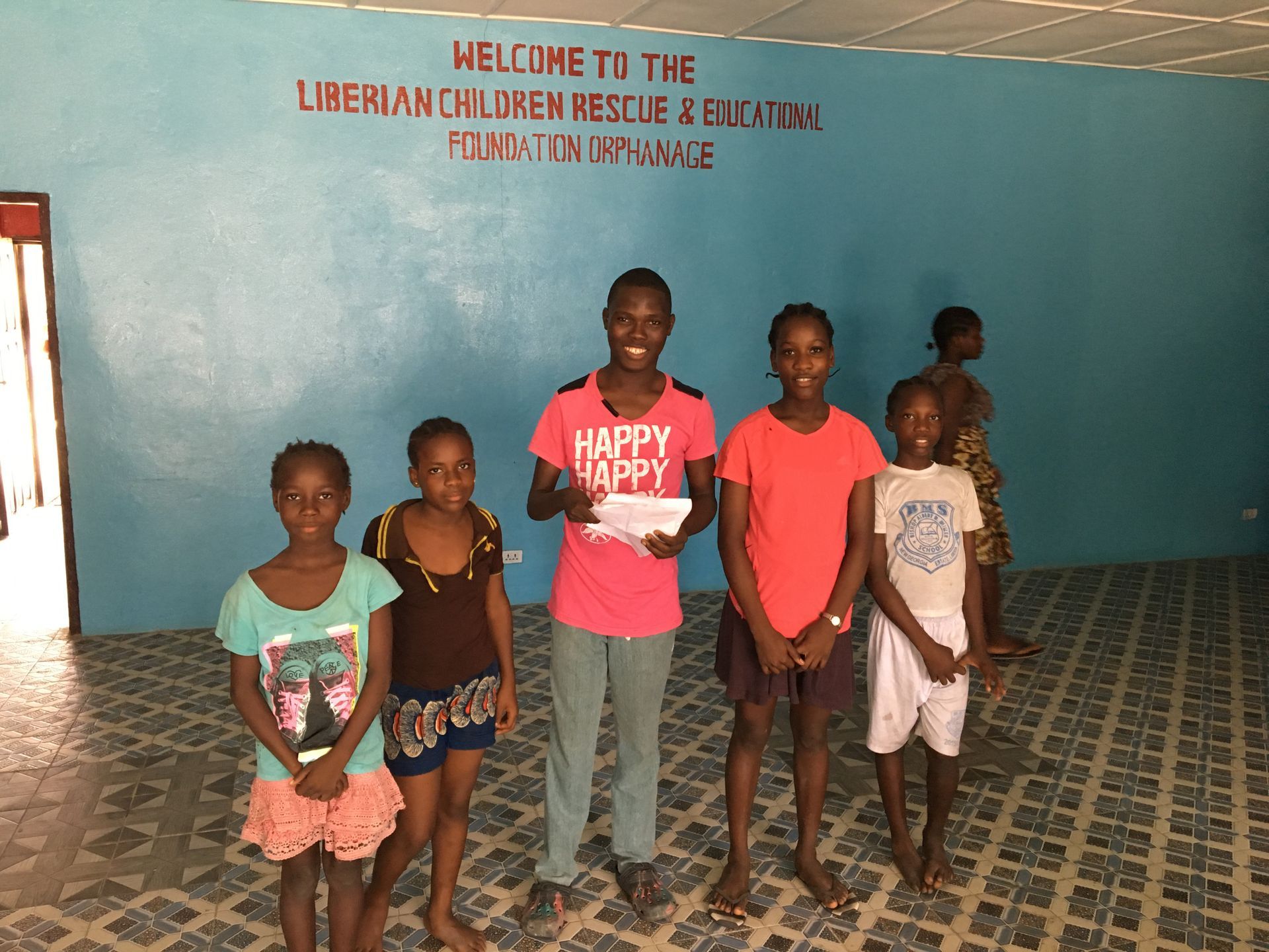 Six children stand in a blue-walled room with a welcome sign on the wall behind them.