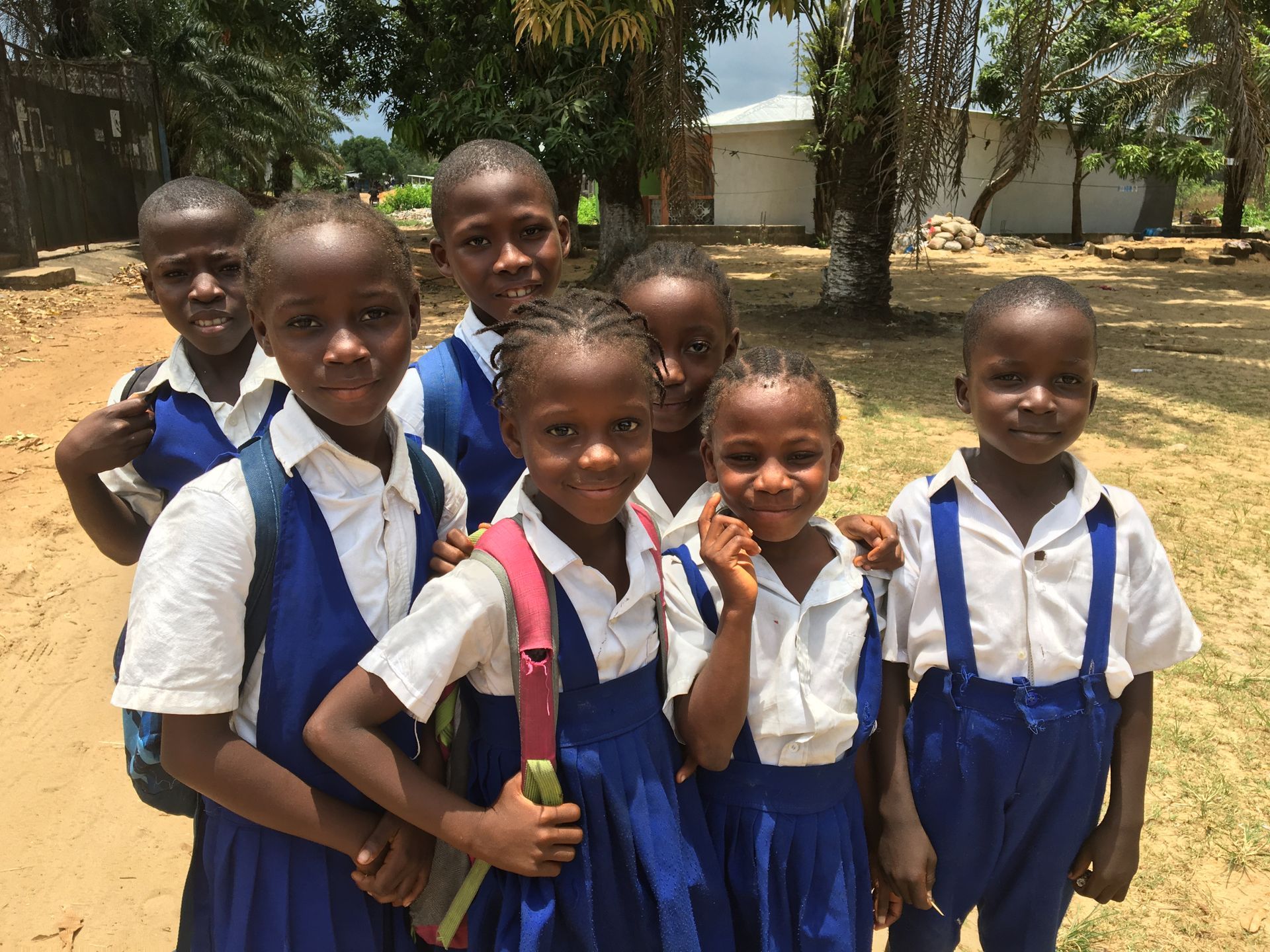 Six schoolchildren in blue uniforms standing outdoors, smiling in a sunny courtyard.