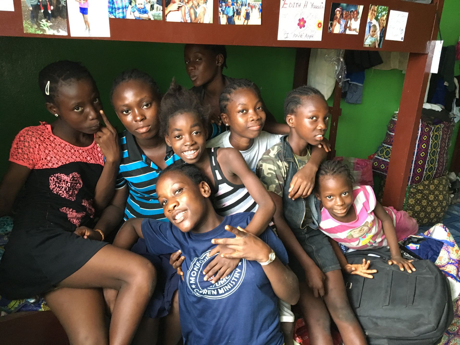 Group of children sitting closely together indoors, smiling and posing on a bed with colorful bags nearby