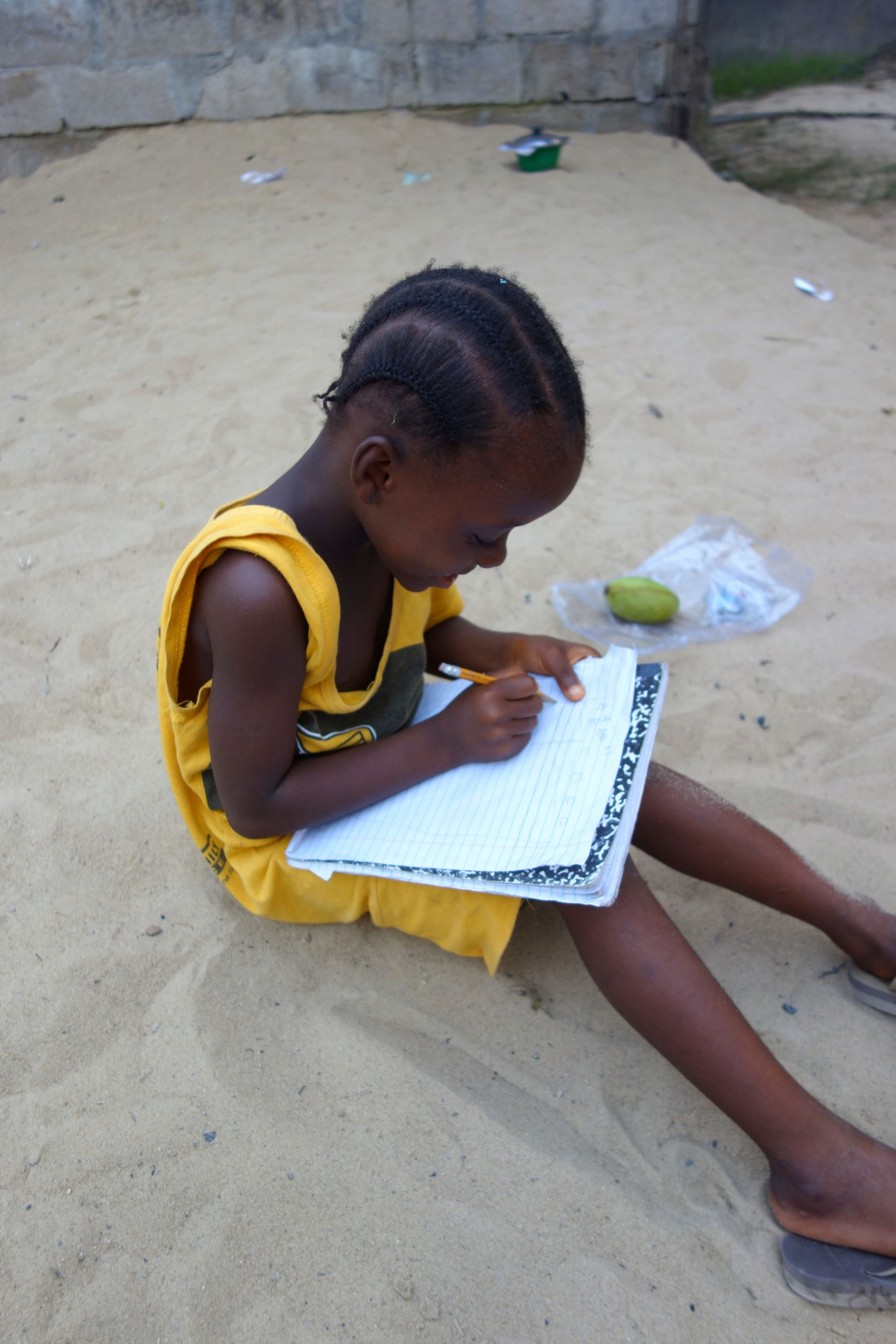 Child in a yellow dress sitting on sand, writing in a notebook with a pencil.