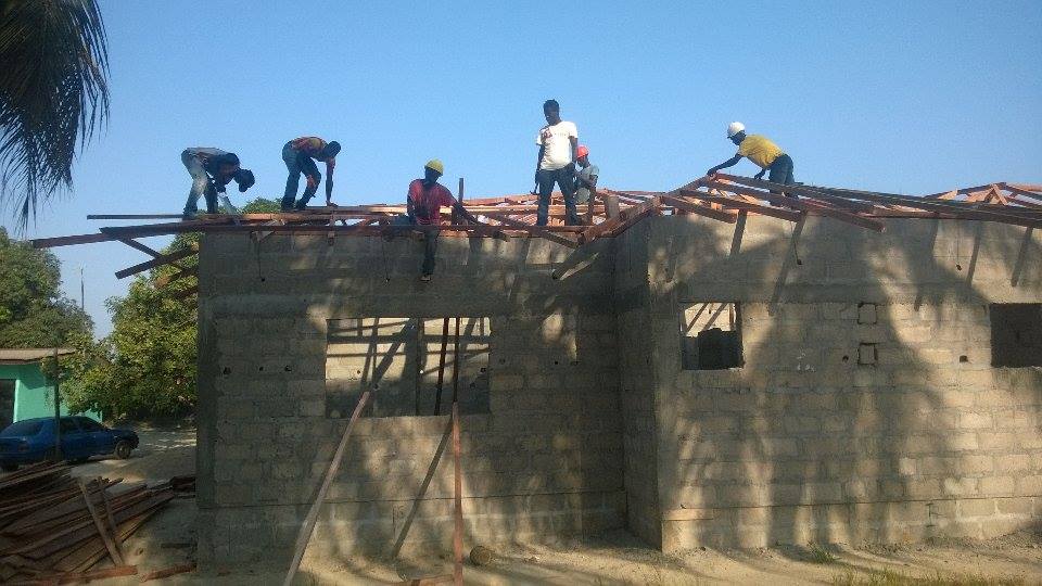 Workers installing a roof on a building under construction, standing on wooden beams against a blue sky.