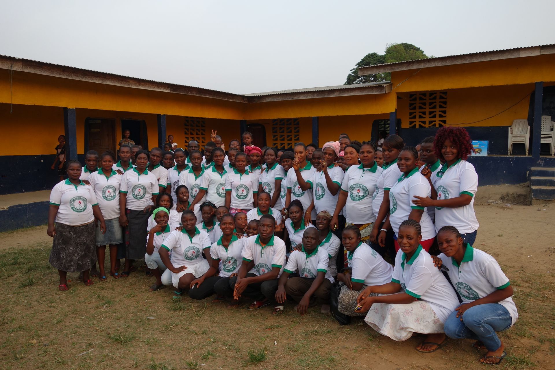 Group posing outdoors in front of a yellow school building, wearing white shirts with green trim.