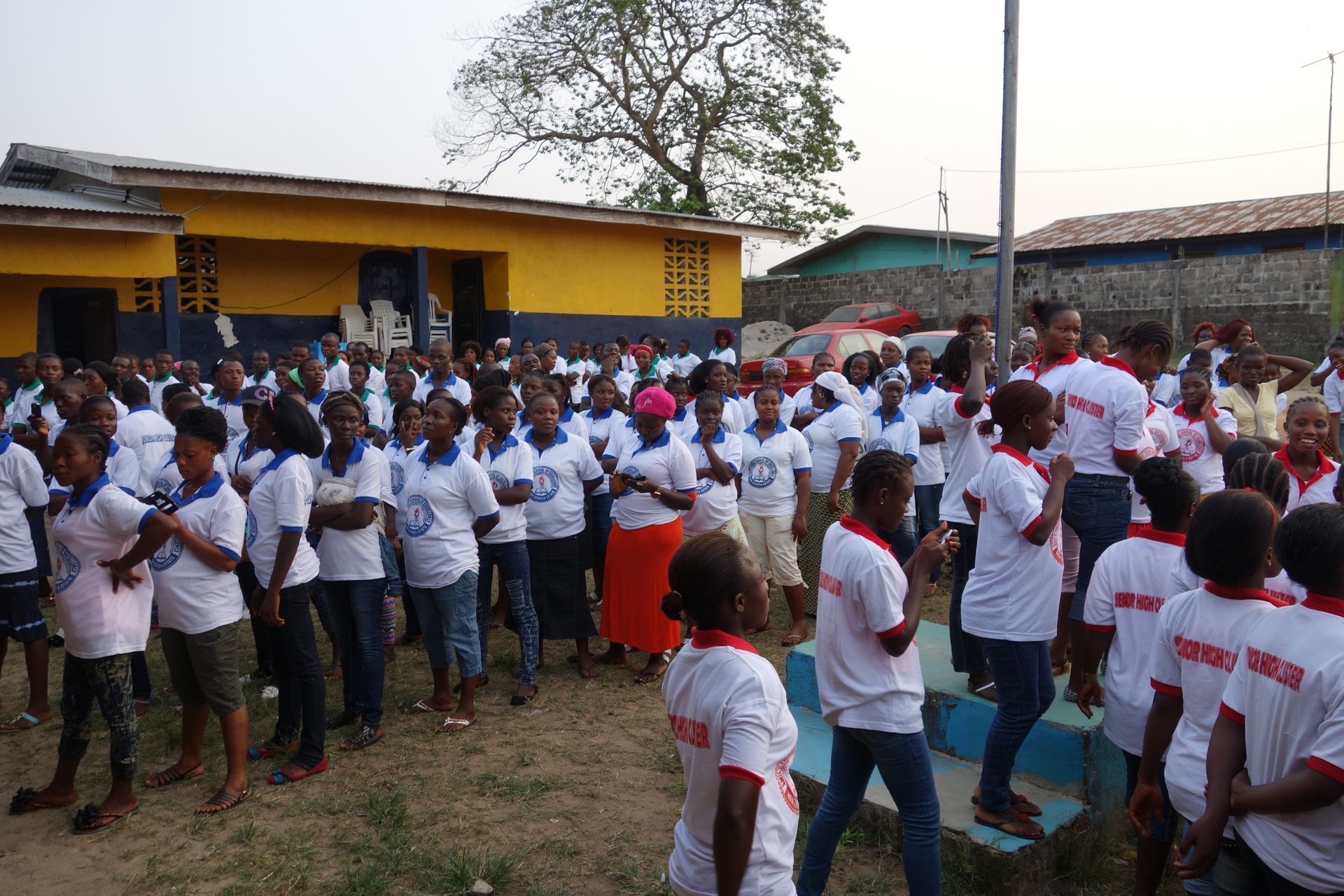 Crowd of people in white shirts gathered outdoors near a yellow building, many clapping and facing forward