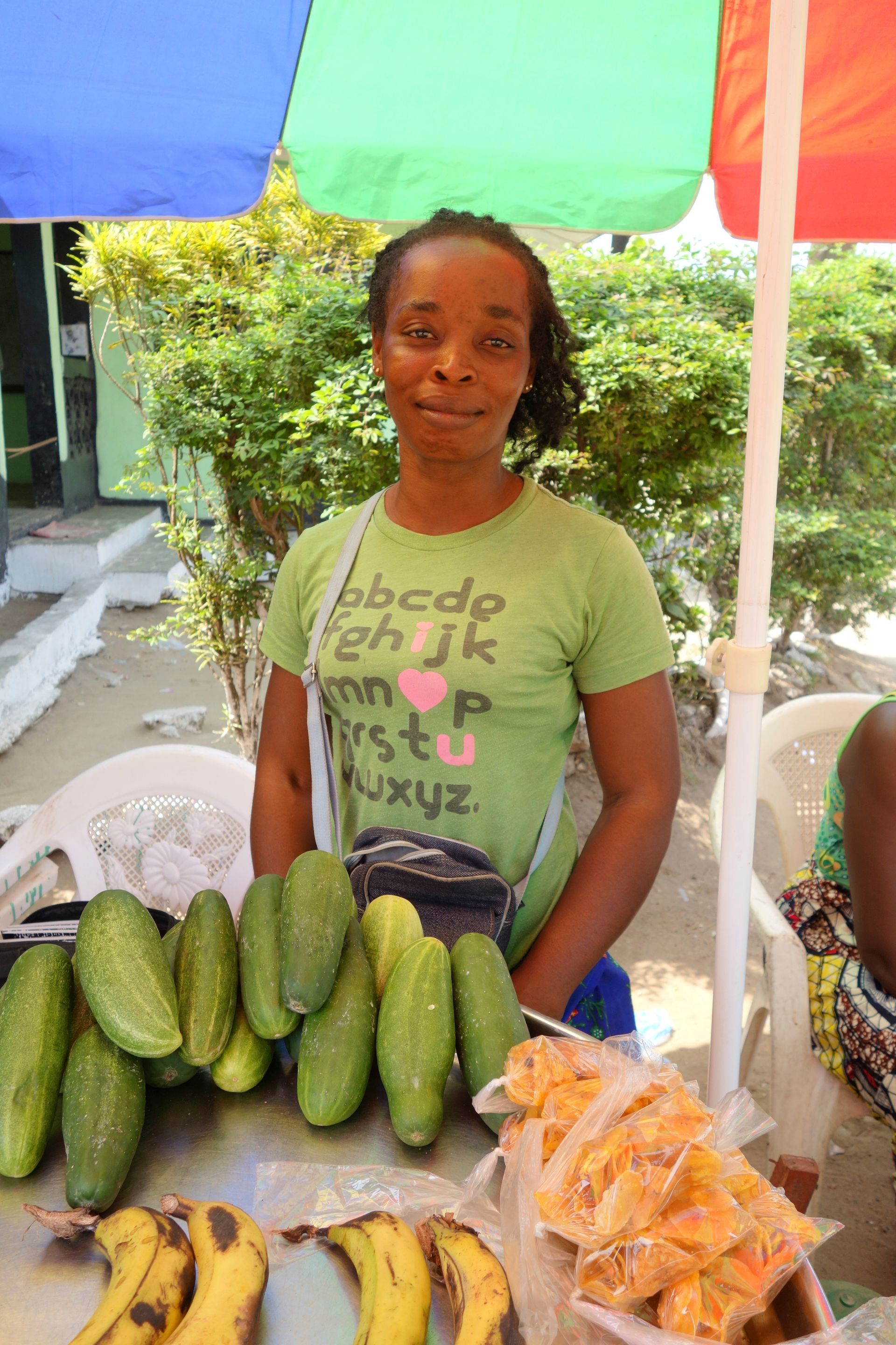 Vendor at a market stall with cucumbers, bananas, and packaged snacks under a colorful umbrella.